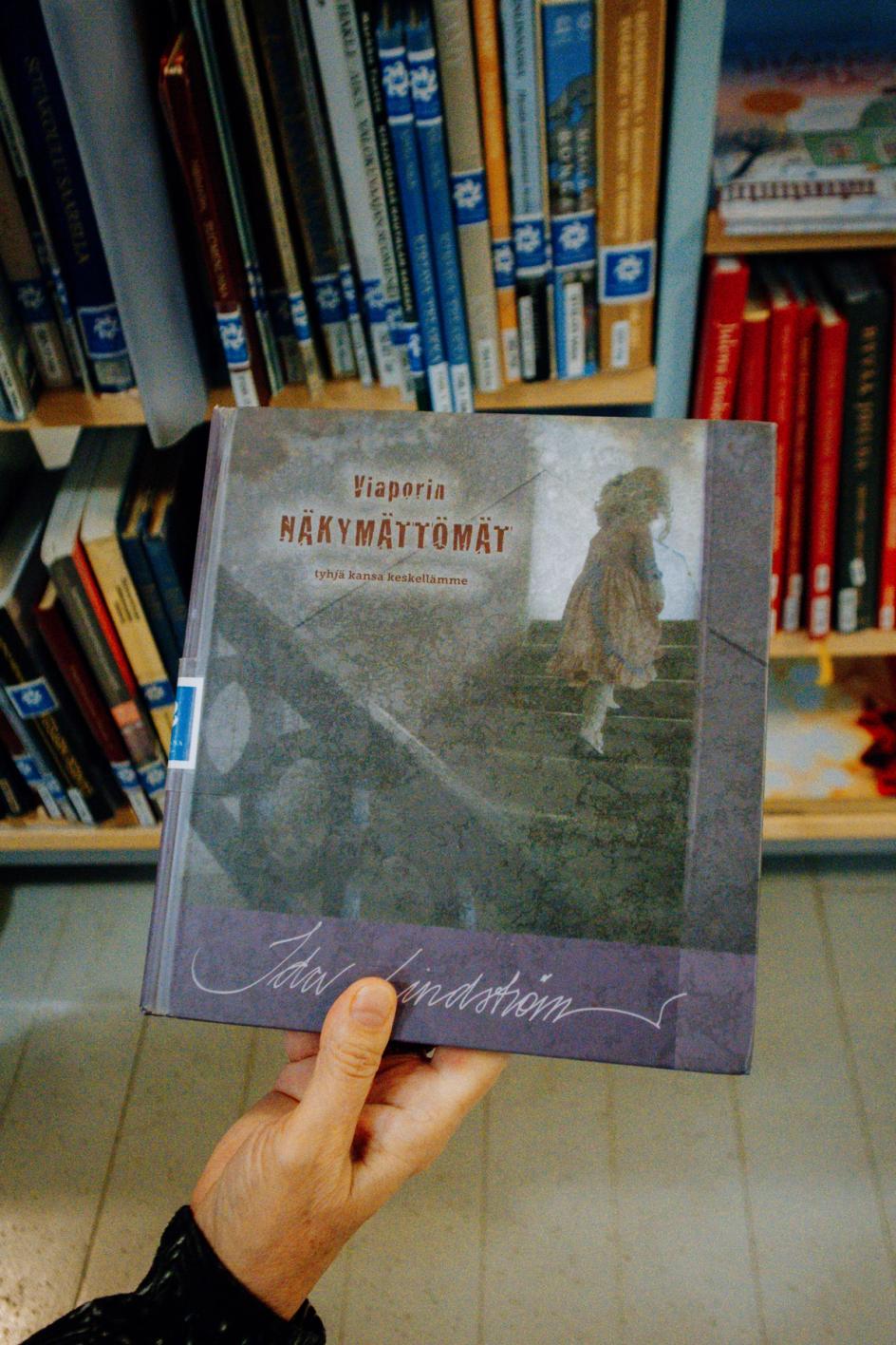 Рука держит книгу Иды Lindström «Viaporin näkymättömät» на фоне библиотечной полки. (Käsi pitelee Ida Lindströmin kirjaa ”Viaporin näkymättömät” kirjaston hyllyn edessä.)