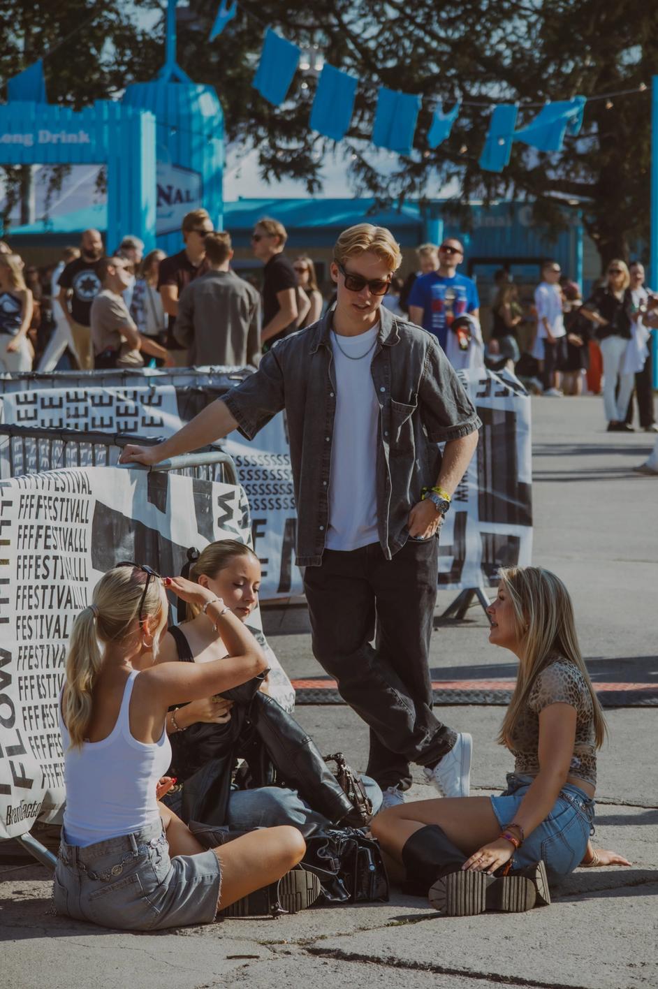 Компания молодых людей на фестивале, трое сидят на земле, один стоит рядом (Nuoria festivaalilla, kolme istuu maassa ja yksi seisoo).