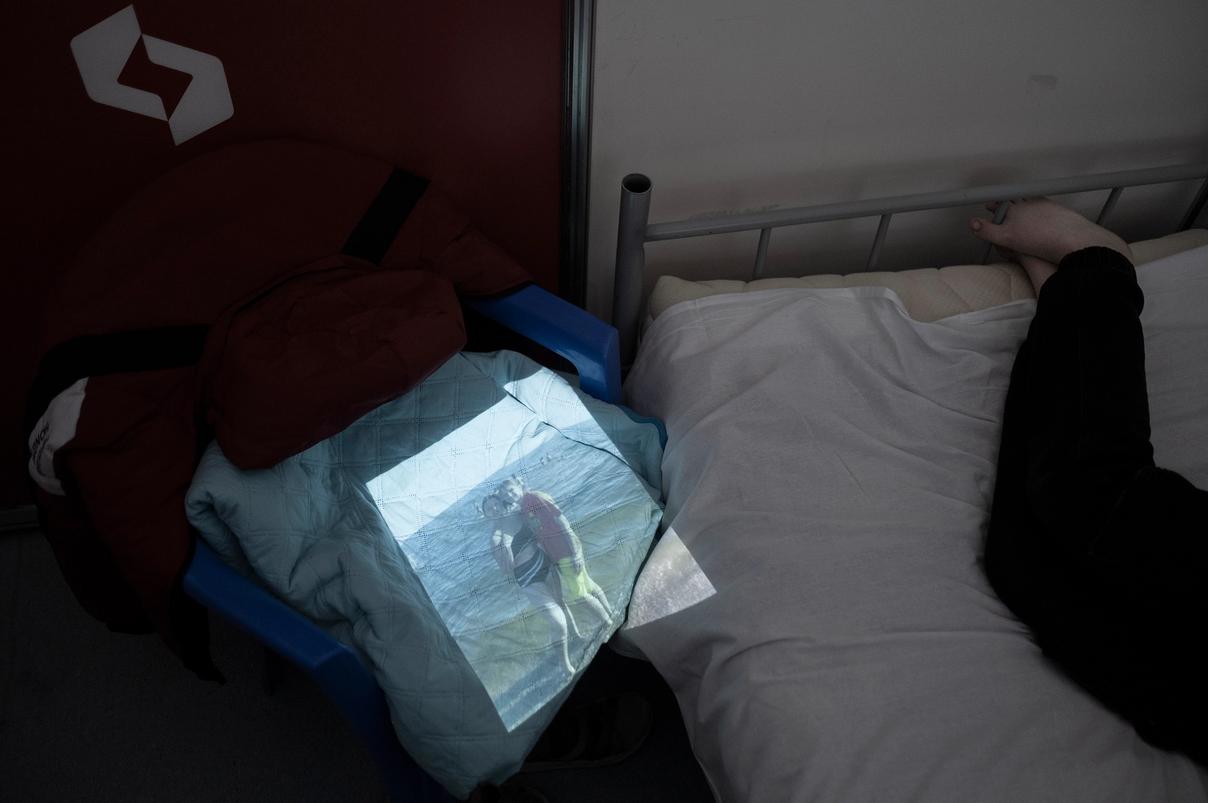 Человек лежит на кровати, рядом на стуле с одеждой проецируется фотография пары на берегу моря (Henkilö makaa sängyllä, vieressä tuolille heijastuu valokuva pariskunnasta meren rannalla)