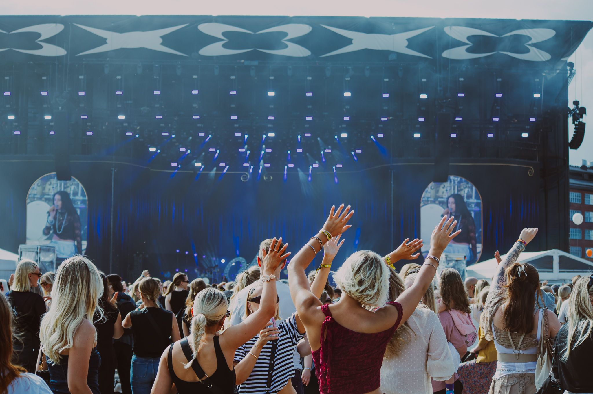 Толпа на фестивале поднимает руки к большой сцене с выступающей артисткой (Yleisö nostaa käsiä suurella festivaalilavalla esiintyjälle).