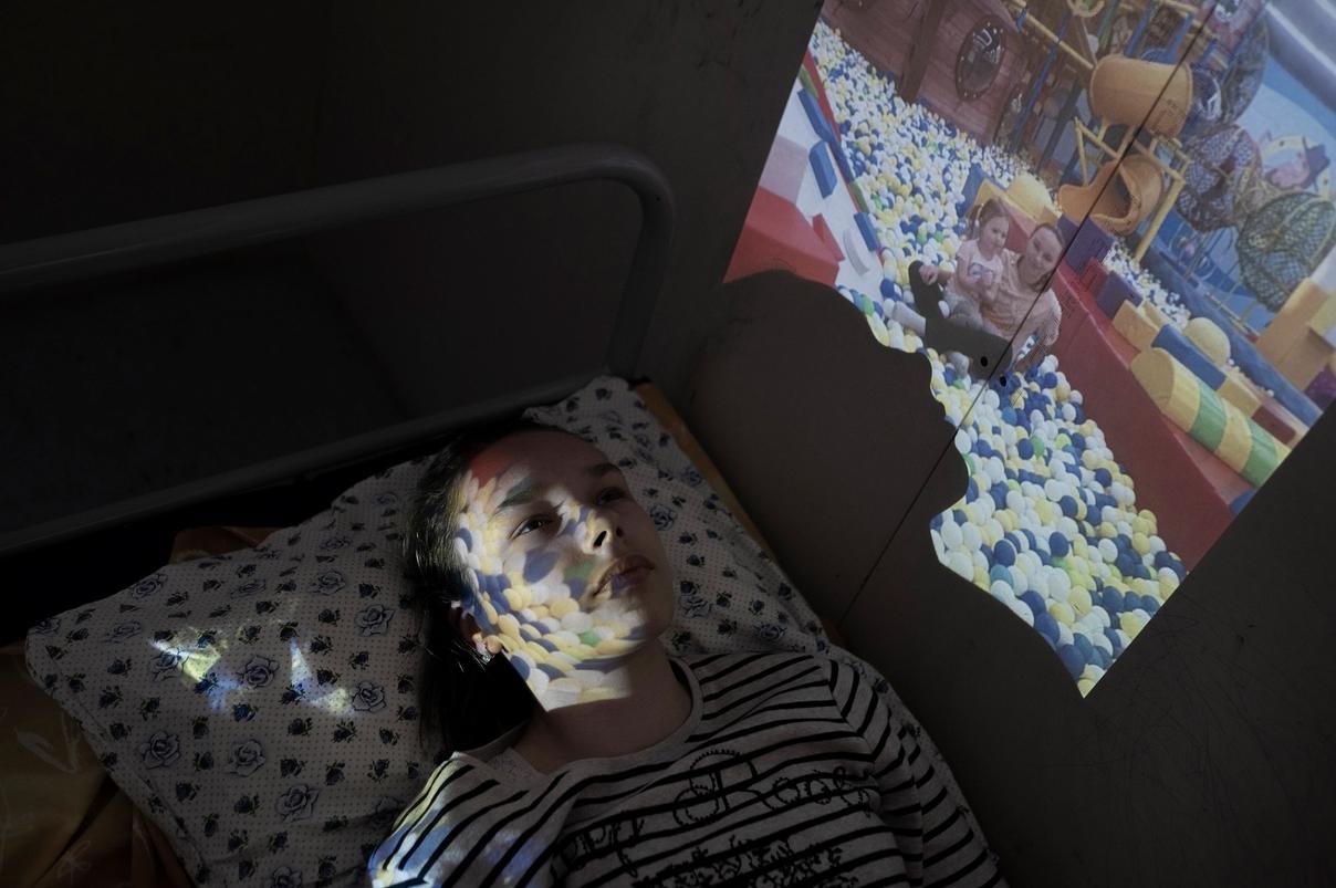 Девочка лежит на кровати, на её лицо проецируется изображение детей в бассейне с шариками (Tyttö makaa sängyllä, hänen kasvoilleen heijastuu kuva lapsista pallomeressä)