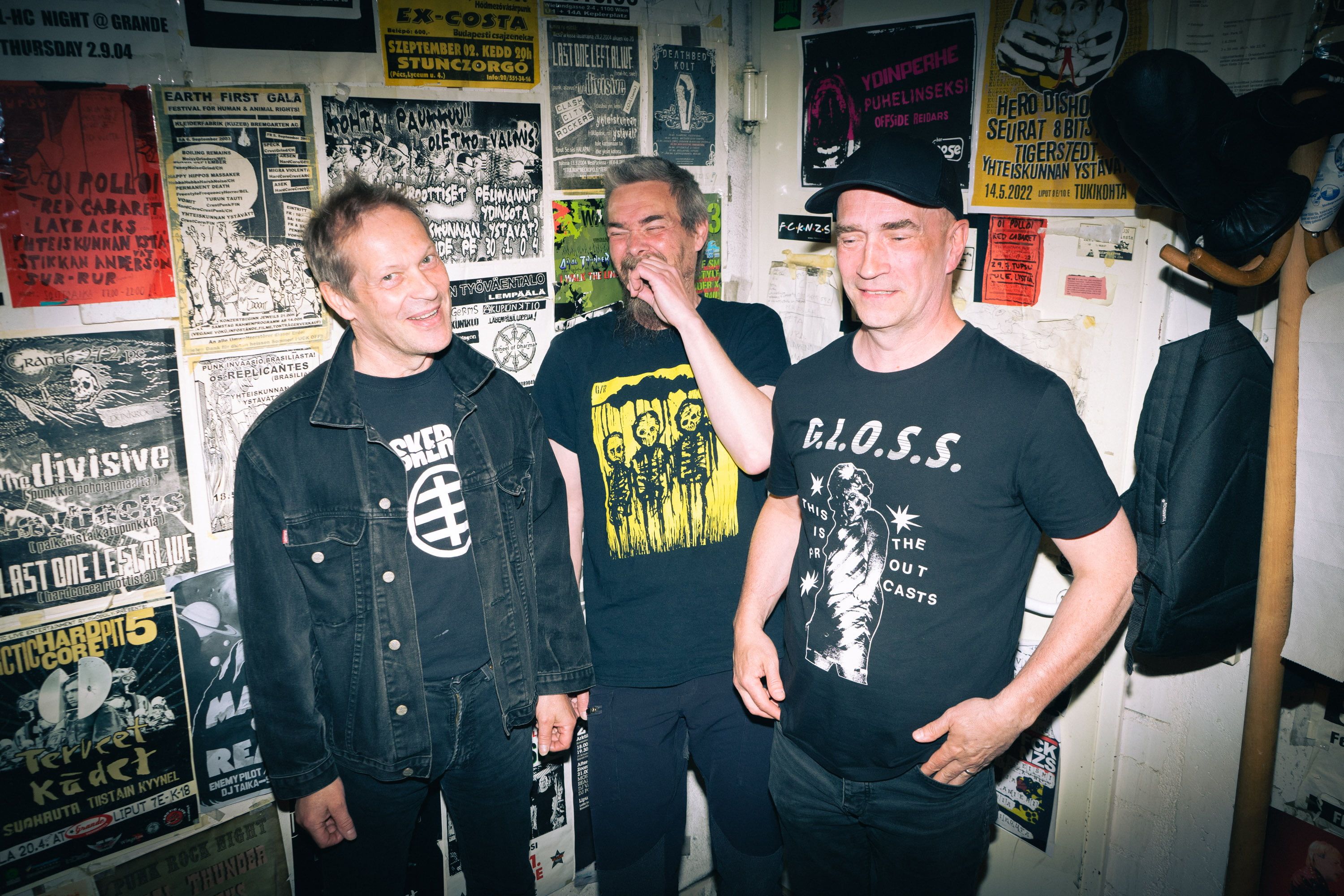 Три улыбающихся панка на фоне стены с плакатами (Kolme hymyilevää punkkaria taustallaan seinä jossa julisteita)