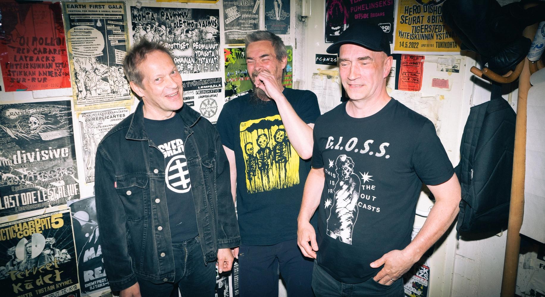 Три улыбающихся панка на фоне стены с плакатами (Kolme hymyilevää punkkaria taustallaan seinä jossa julisteita)