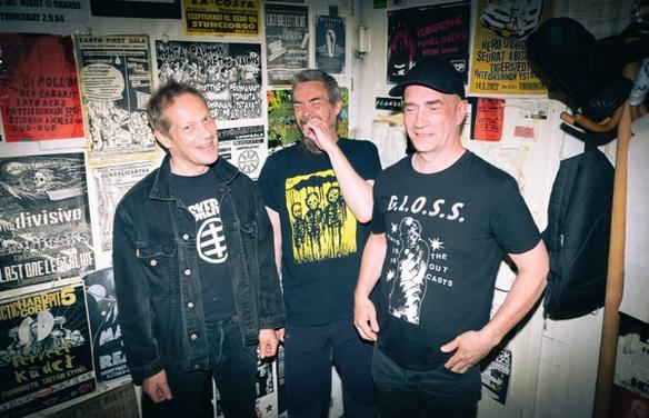 Три улыбающихся панка на фоне стены с плакатами (Kolme hymyilevää punkkaria taustallaan seinä jossa julisteita)