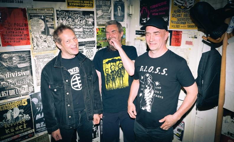 Три улыбающихся панка на фоне стены с плакатами (Kolme hymyilevää punkkaria taustallaan seinä jossa julisteita)