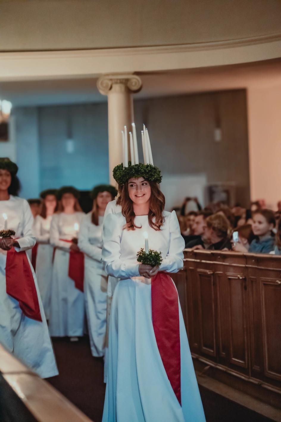 Vuoden 2023 Lucia-neito Madeleine Amoroso kävelee hymyillen kulkue perässään, kynttiläkruunu päässään.