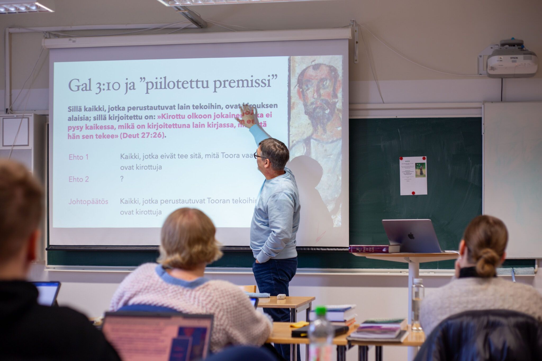 Лектор указывает на презентацию о «скрытой посылке» (Luennoitsija osoittaa esitystä)