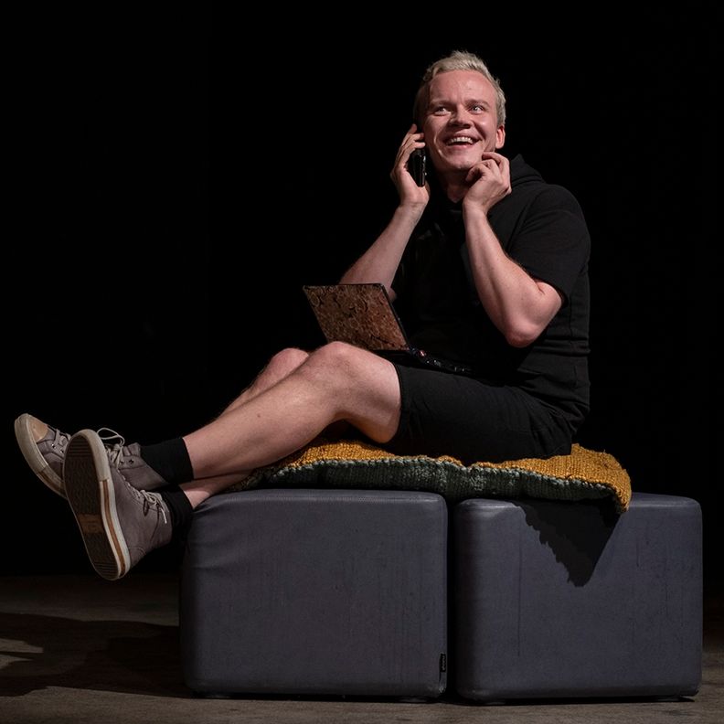 Мужчина на сцене сидит на пуфе, говорит по телефону (Mies istuu lavalla rahilla ja puhuu puhelimeen)