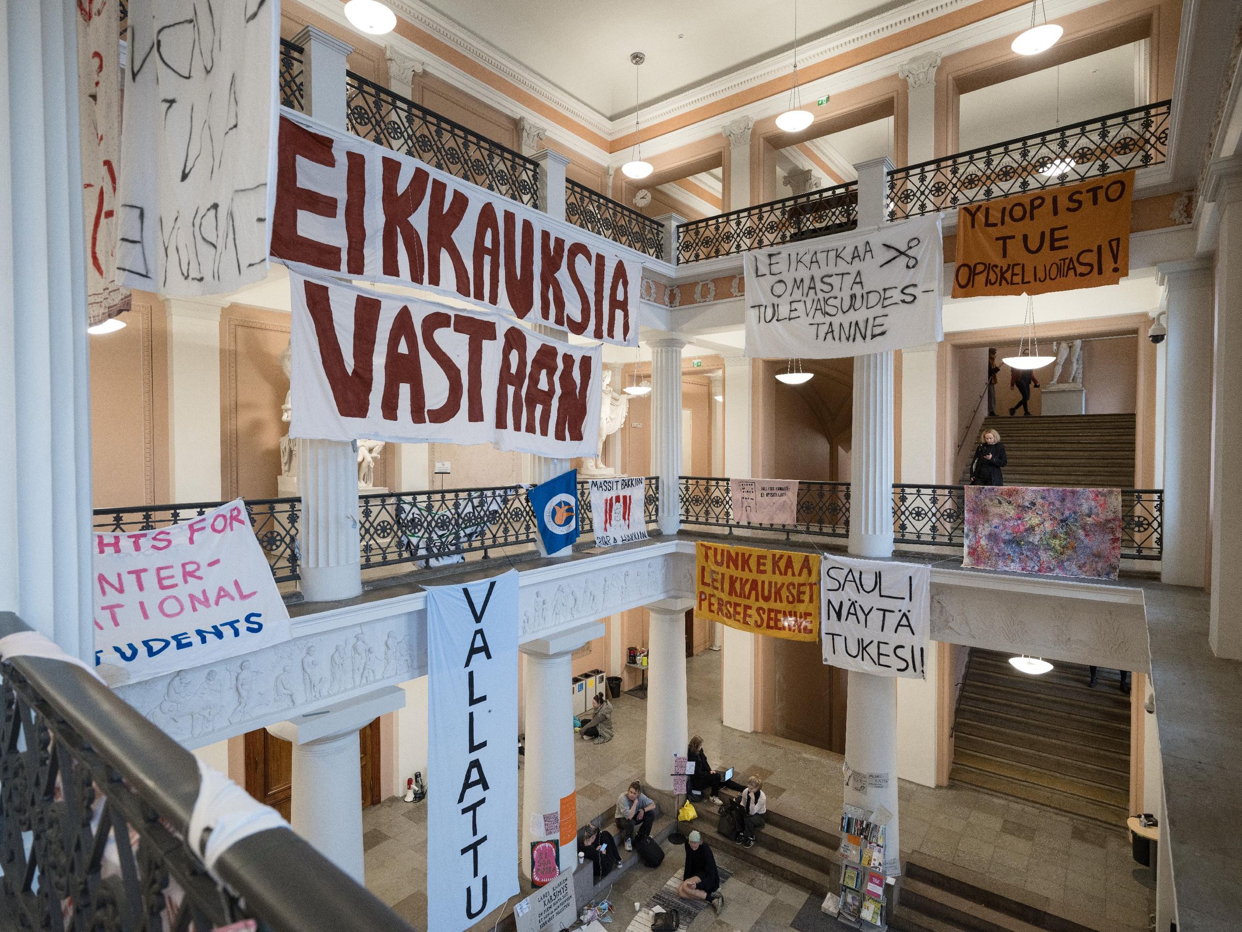 Näkymä Helsingin yliopiston päärakennuksen aulasta, jonne opiskelijat ovat asetelleet banderolleja protestina Petteri Orpon hallituksen ajamaa leikkauspolitiikkaa vastaan. Suurissa banderolleissa lukee muun muassa "Leikkauksia vastaan" ja "Yliopisto, tue opiskelijoitasi!". Aula- ja porrastiloissa istuu opiskelijoita lattialla, osa työskentelee tietokoneilla.