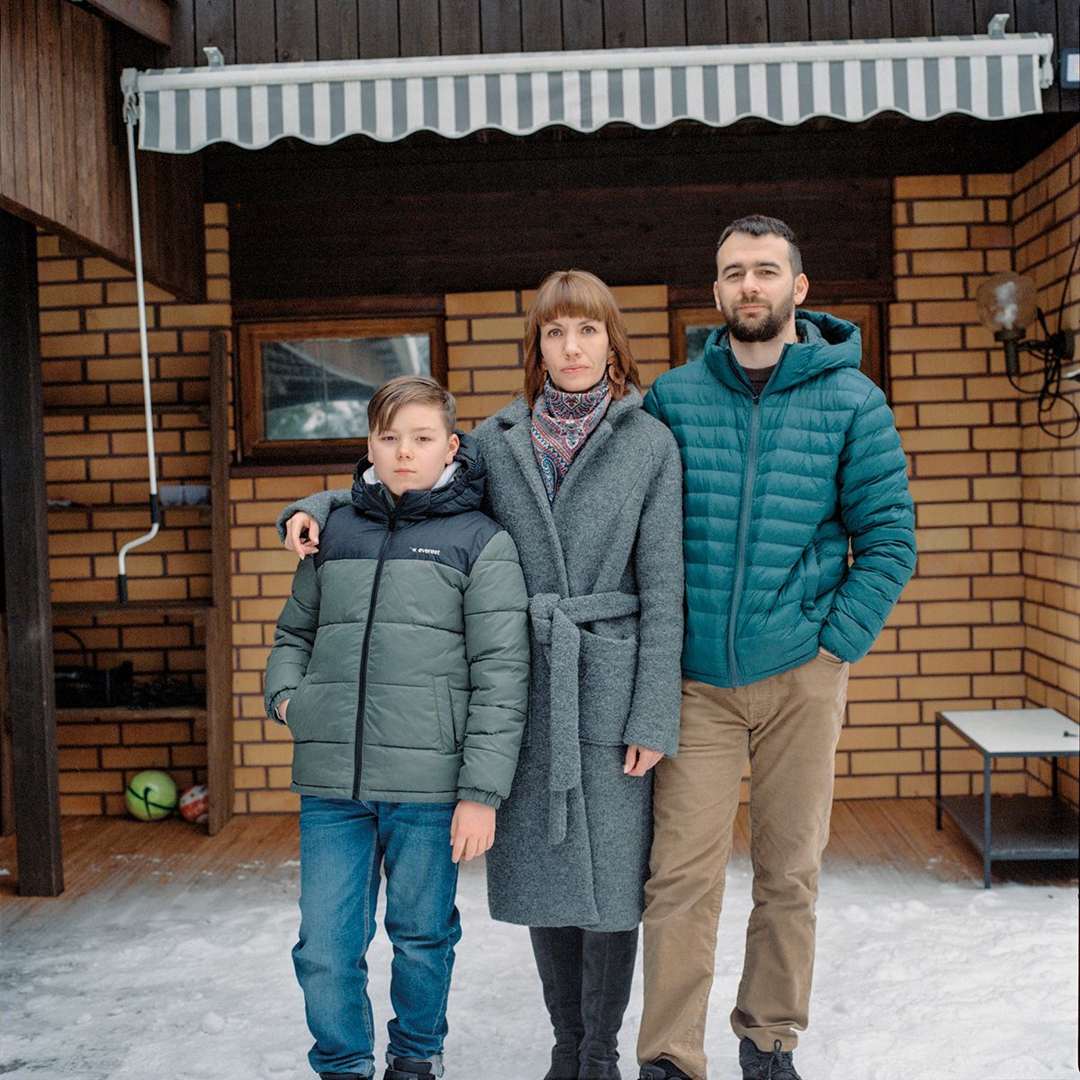 Семья из трёх человек стоит на улице у дома зимой (Kolmihenkinen perhe seisoo talon edessä talvella).
