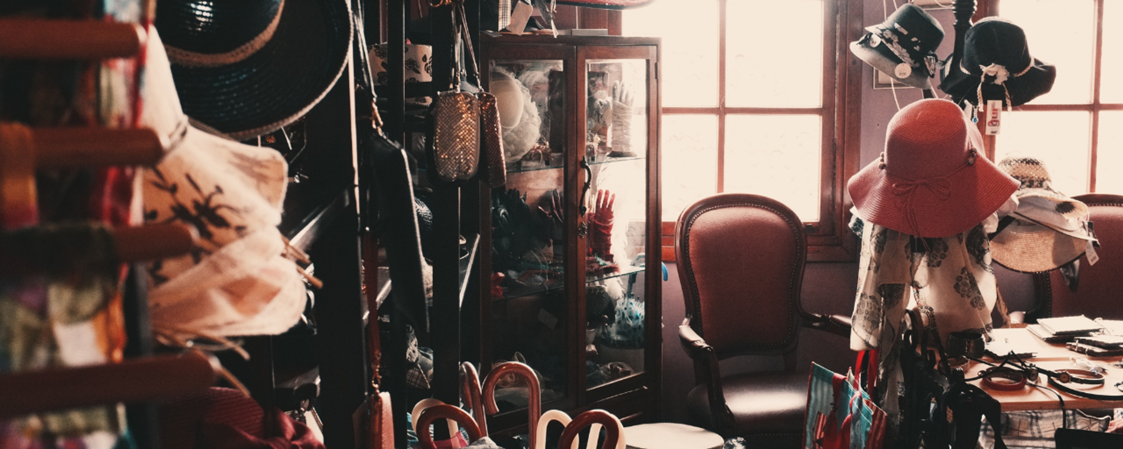 A cluttered, vintage-style shop interior filled with various hats, gloves in glass display cases, and antique chairs bathed in soft natural light from a window.