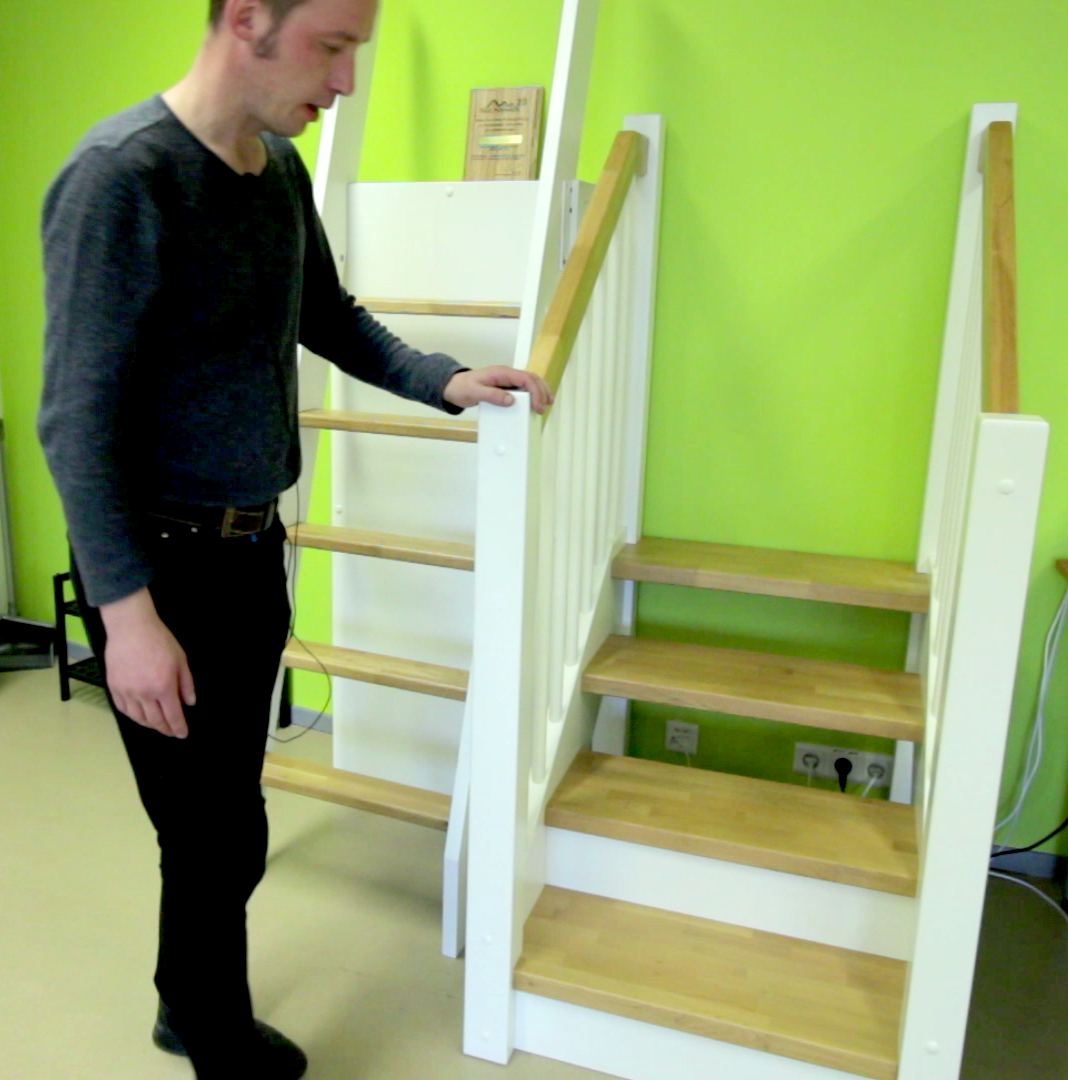 A man stands next to a display comparing two types of wooden stairs: a steep, space-saving attic ladder and a standard staircase with white-painted stringers and light-wood treads.