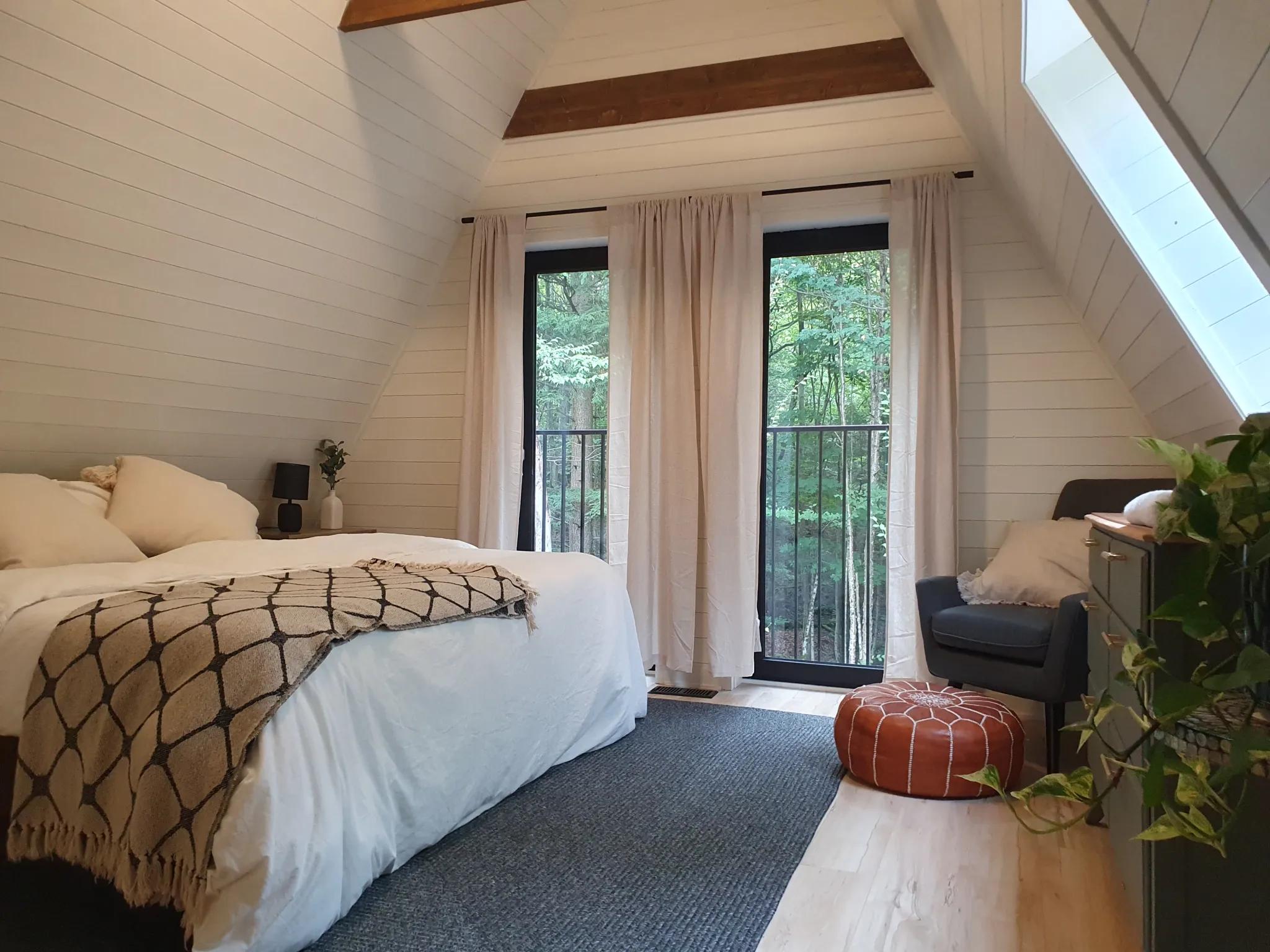 A cozy bedroom in an A-frame loft features white shiplap walls, a neutral bed set with a patterned throw, and tall windows that overlook a lush green forest.