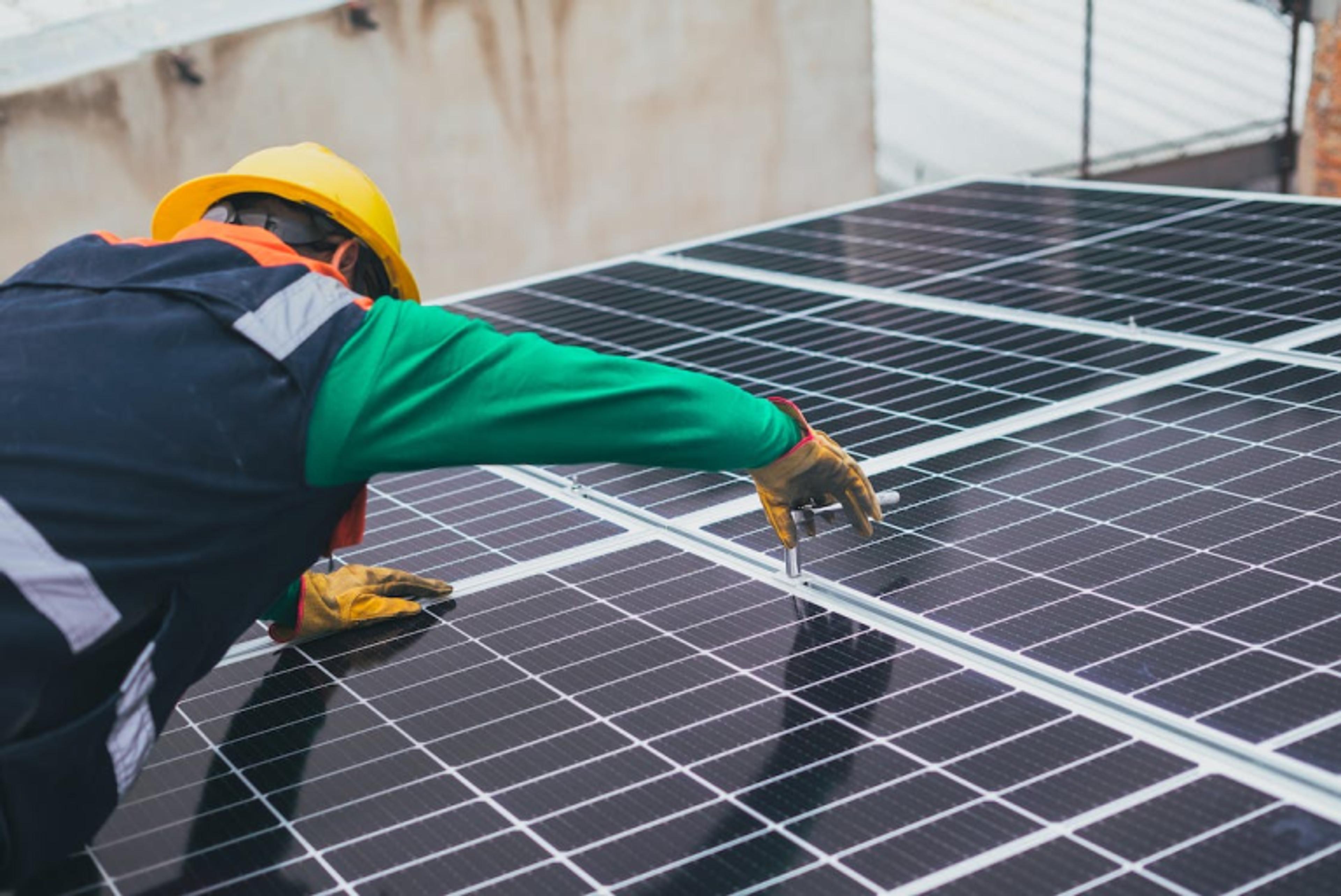 A technician in a yellow hard hat and protective gloves uses a tool to secure a solar panel as part of a rooftop installation.