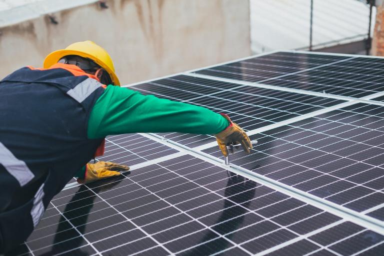 A technician in a yellow hard hat and protective gloves uses a tool to secure a solar panel as part of a rooftop installation.