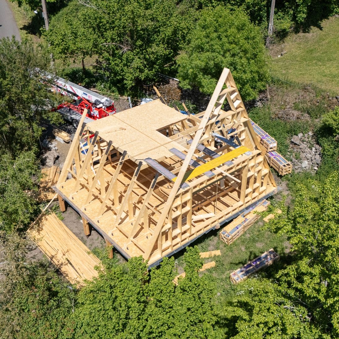 Aerial view of an Avrame Trio 75 A-frame kit under active assembly in France, showing the timber frame structure and roof trusses with a crane nearby