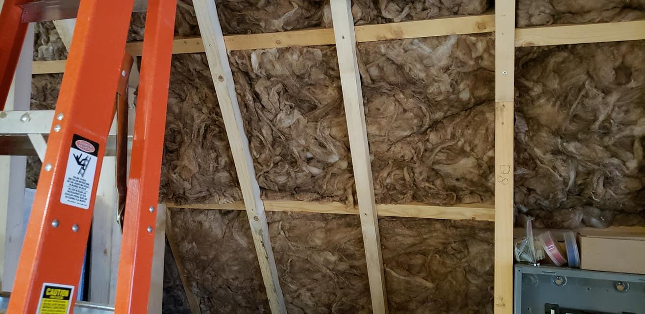 Batts of brown fiberglass insulation are tucked into the wooden wall studs of a building under construction, with an orange ladder visible in the foreground.