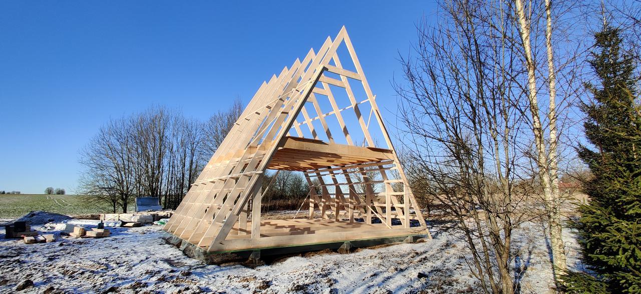 The skeleton of a wooden A-frame house stands in a snowy field against a clear blue sky, showing the progress of the structural trusses and flooring.