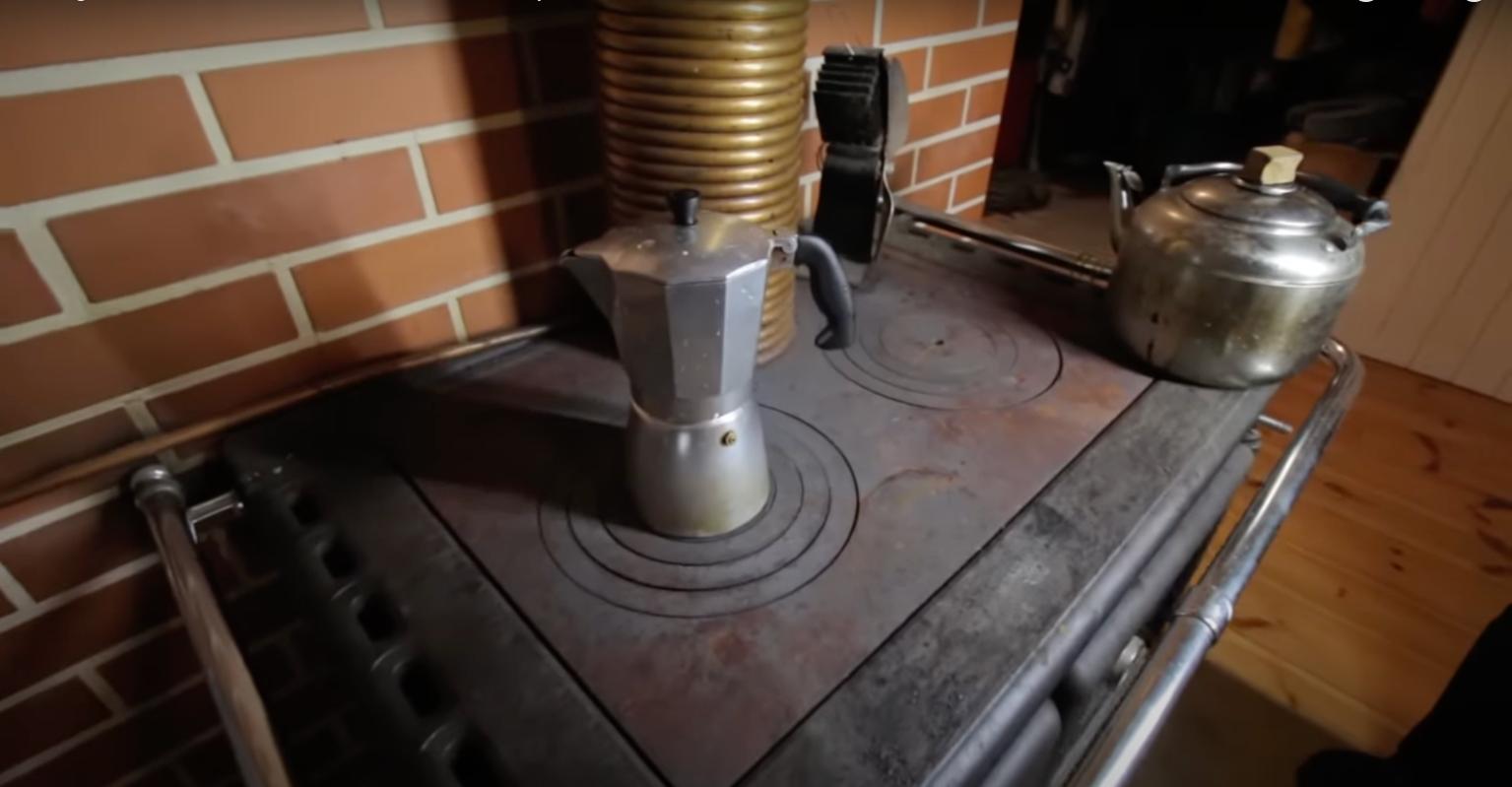 An aluminum moka pot and a vintage metal tea kettle sit atop a traditional wood-burning cast-iron stove against a brick wall.