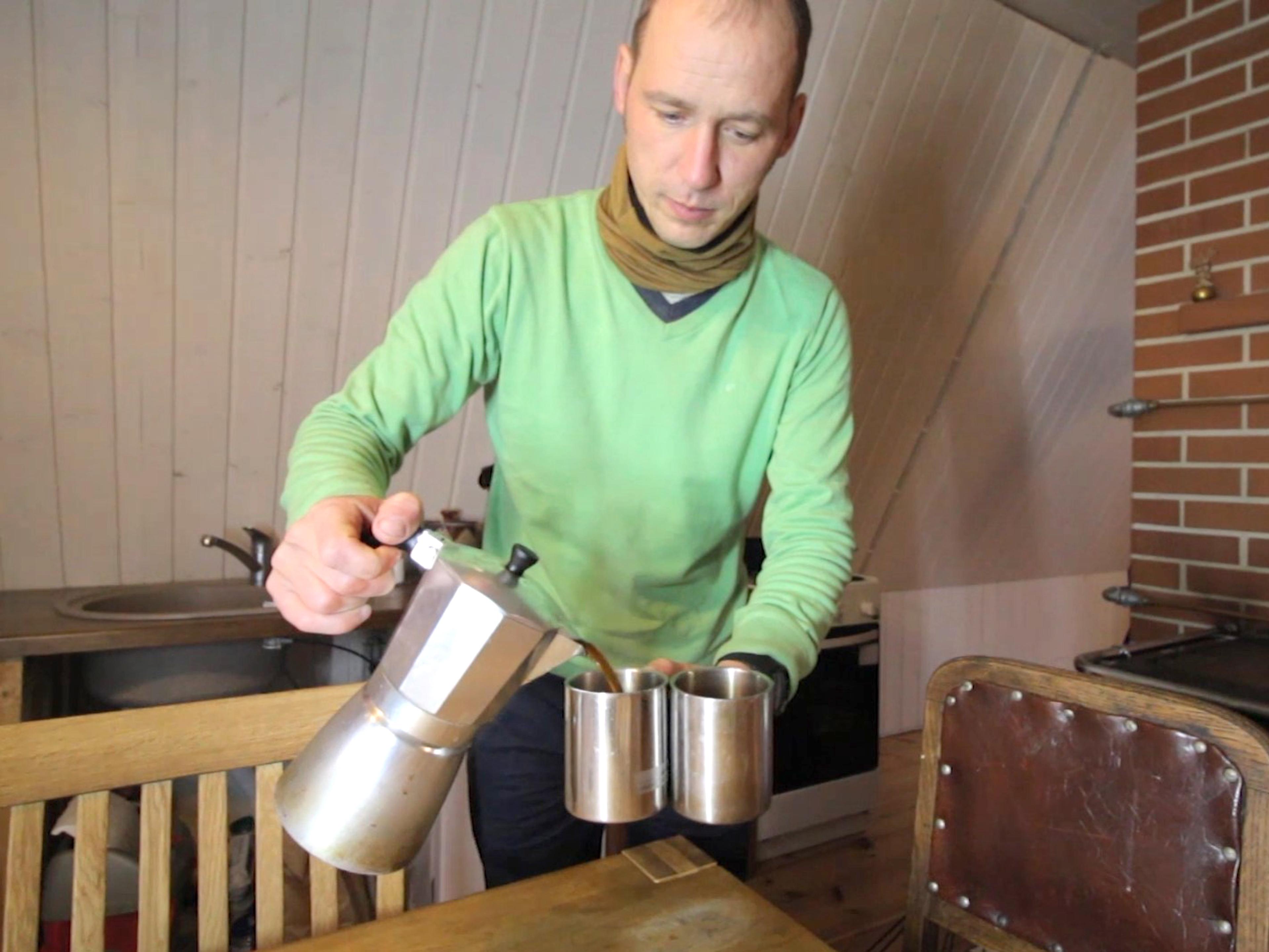 A man in a green sweater pours coffee from a moka pot into two metal mugs inside a rustic cabin.