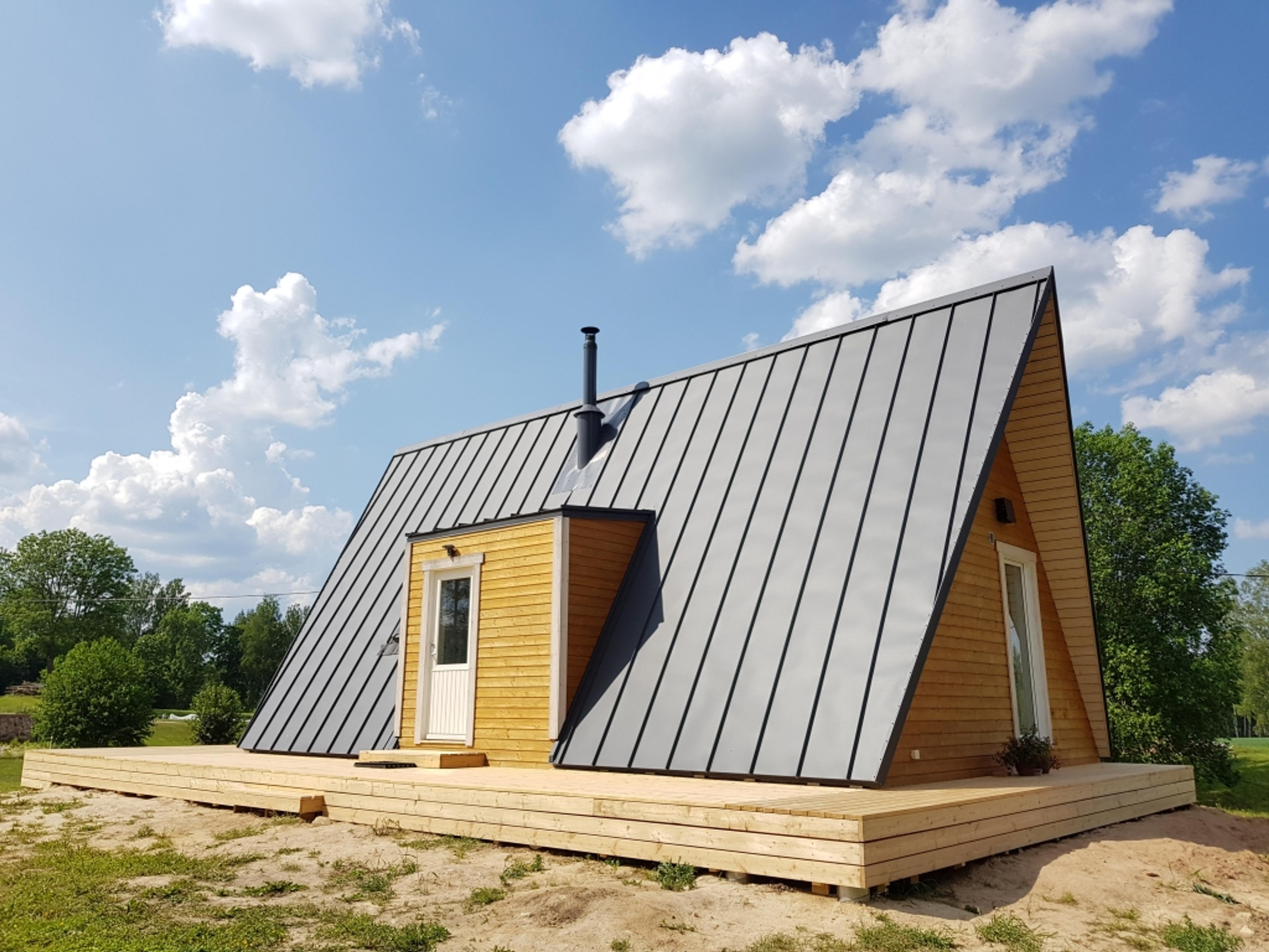 A light-wood A-frame house featuring a gray metal roof and a matching side dormer sits on a large wooden platform under a bright, cloud-filled sky.