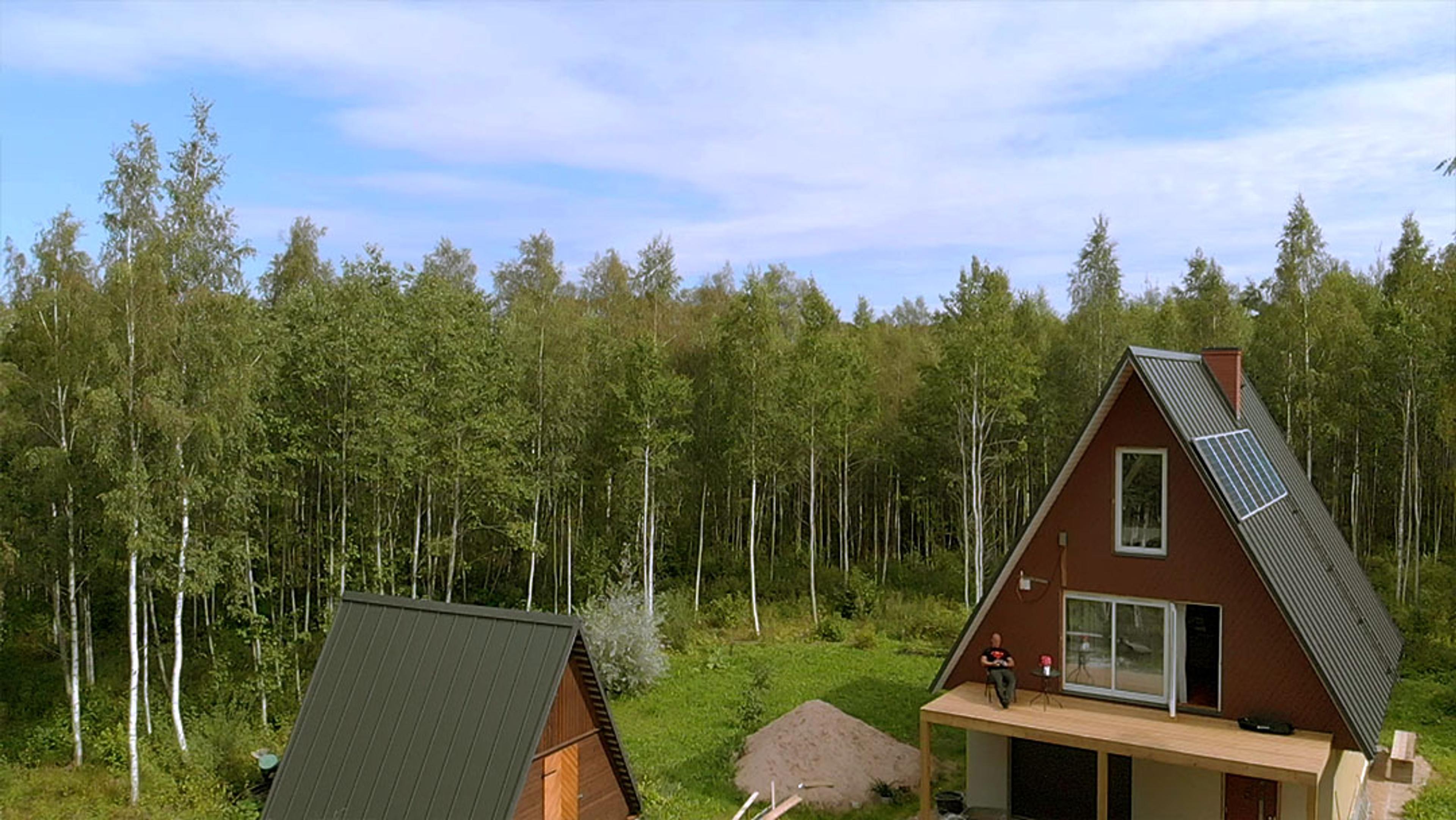 A person sits on the deck of a brown A-frame house with solar panels, situated next to a smaller A-frame outbuilding in a green forest clearing.