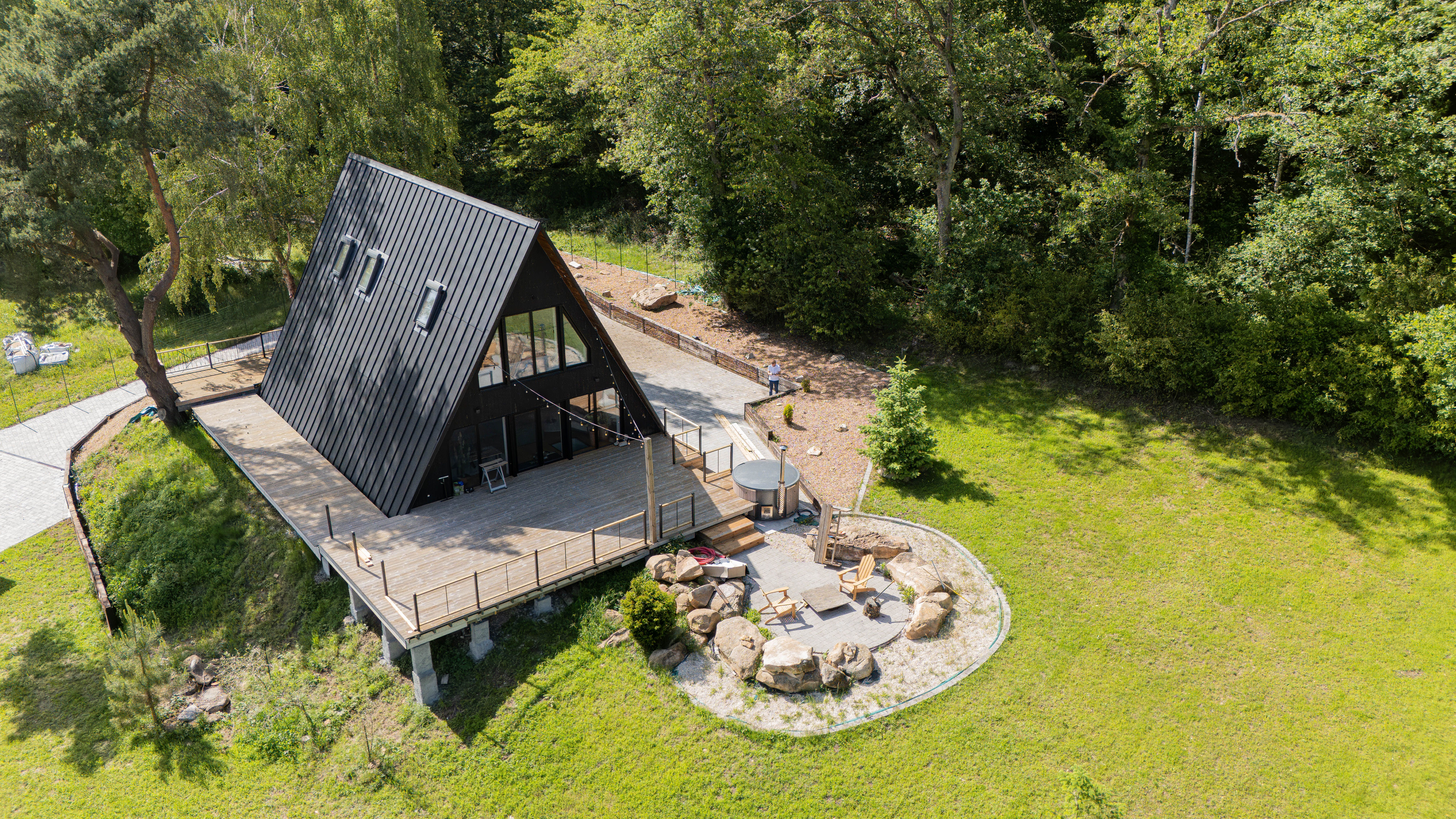 An high-angle, aerial view of a large, modern black A-frame cabin built on a hillside clearing bordered by thick green trees. The cabin features a dark metal standing-seam roof with integrated roof windows, a dark facade with large glass windows, and an expansive elevated wooden deck wrapping around the front. Next to the deck, a circular paved stone patio is designed into the lawn, complete with wooden lounge chairs, rock landscaping features, and an outdoor hot tub.