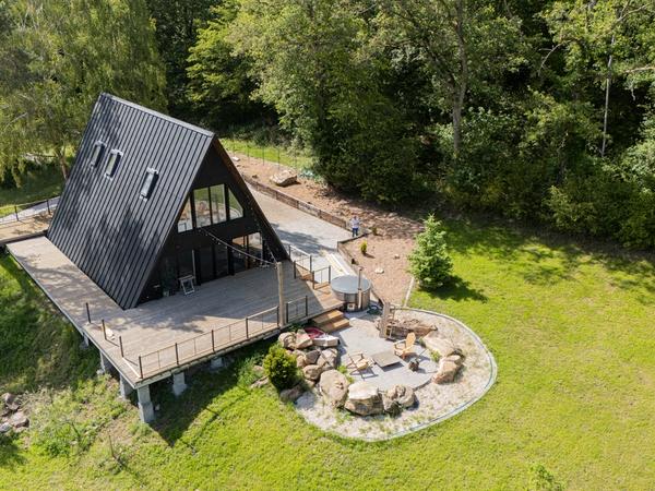 An high-angle, aerial view of a large, modern black A-frame cabin built on a hillside clearing bordered by thick green trees. The cabin features a dark metal standing-seam roof with integrated roof windows, a dark facade with large glass windows, and an expansive elevated wooden deck wrapping around the front. Next to the deck, a circular paved stone patio is designed into the lawn, complete with wooden lounge chairs, rock landscaping features, and an outdoor hot tub.