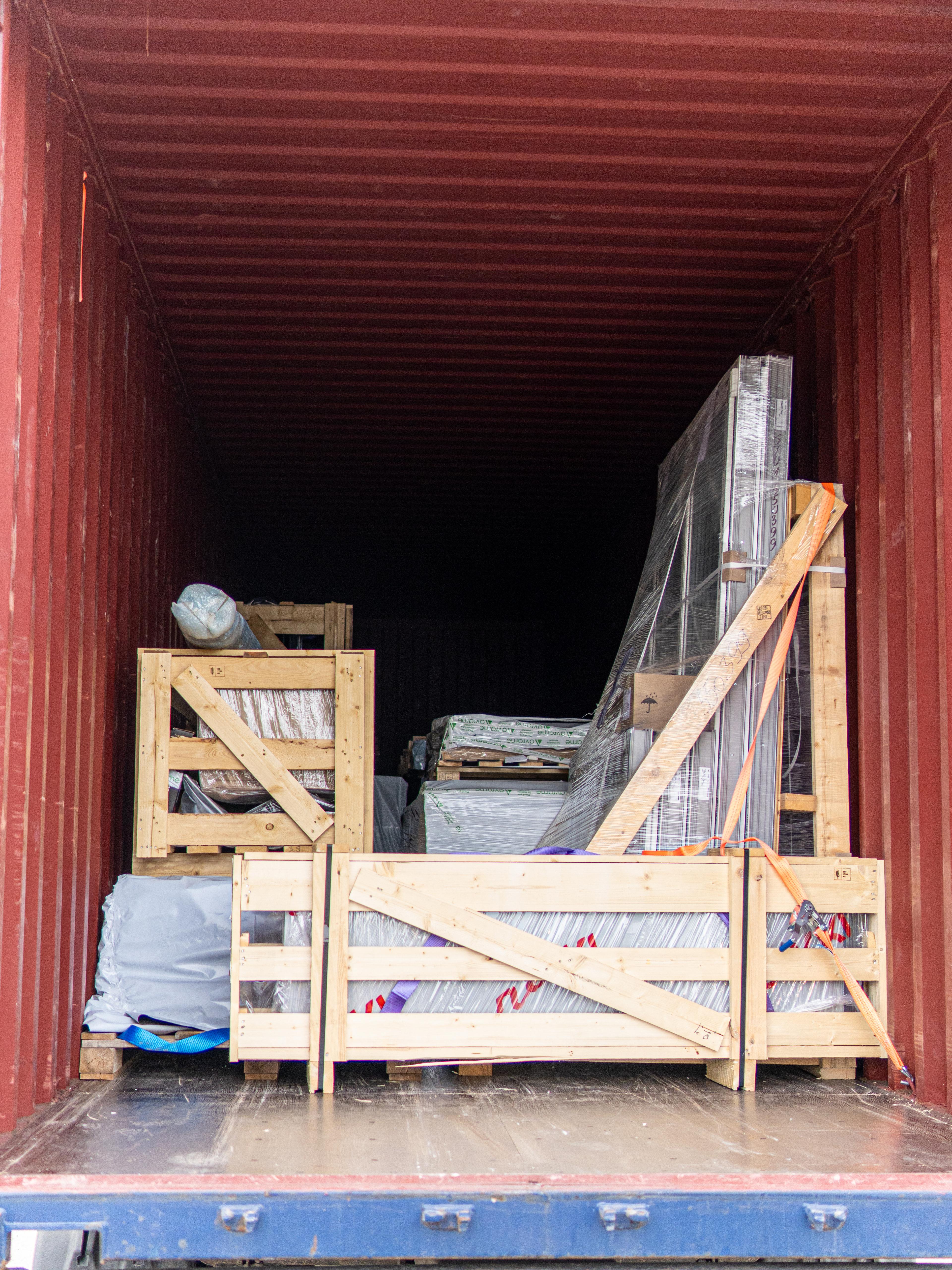 he open doors of a large red shipping container, revealing the neatly packed contents of an Avrame kit home. The interior displays various building materials, including large flat-packed components, window units wrapped in protective plastic, and structural elements secured within custom-built wooden crates for safe transport.