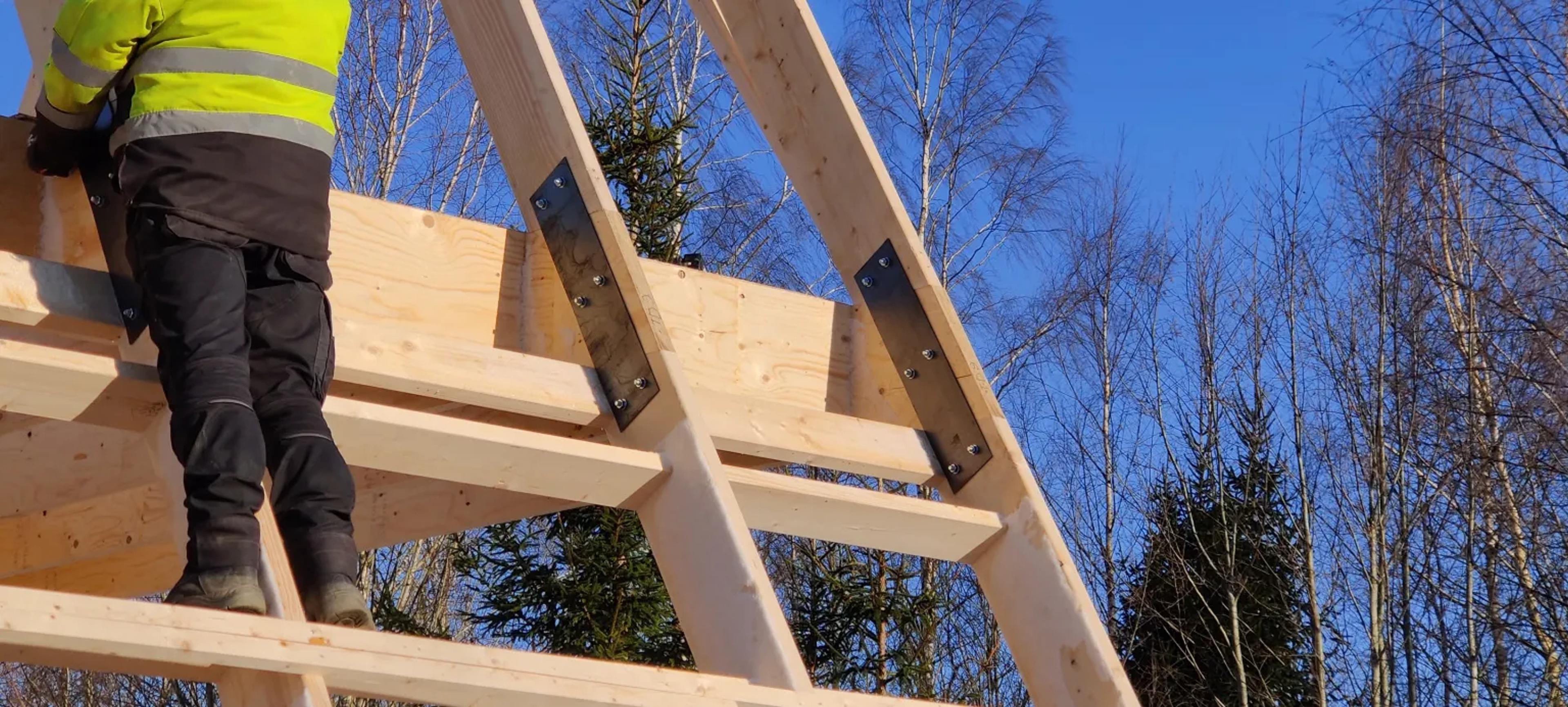 A construction worker in a high-visibility jacket stands on the wooden framework of an A-frame building, where metal brackets secure the rafters under a clear blue sky.