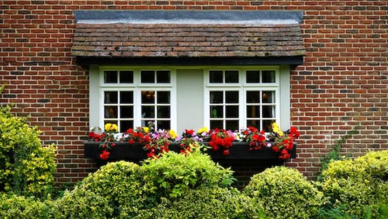 Two white-paned windows are set into a red brick wall and accented with a dark window box filled with vibrant red and yellow flowers.