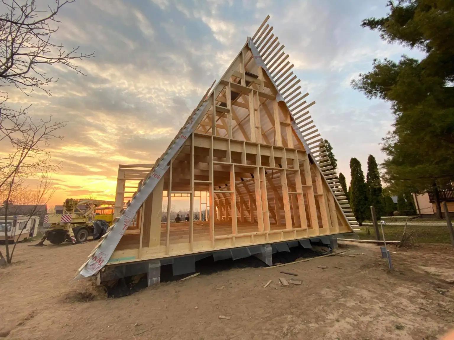 The wooden structural framing of an A-frame house stands on a concrete foundation during a golden sunset, with a construction crane visible in the background.