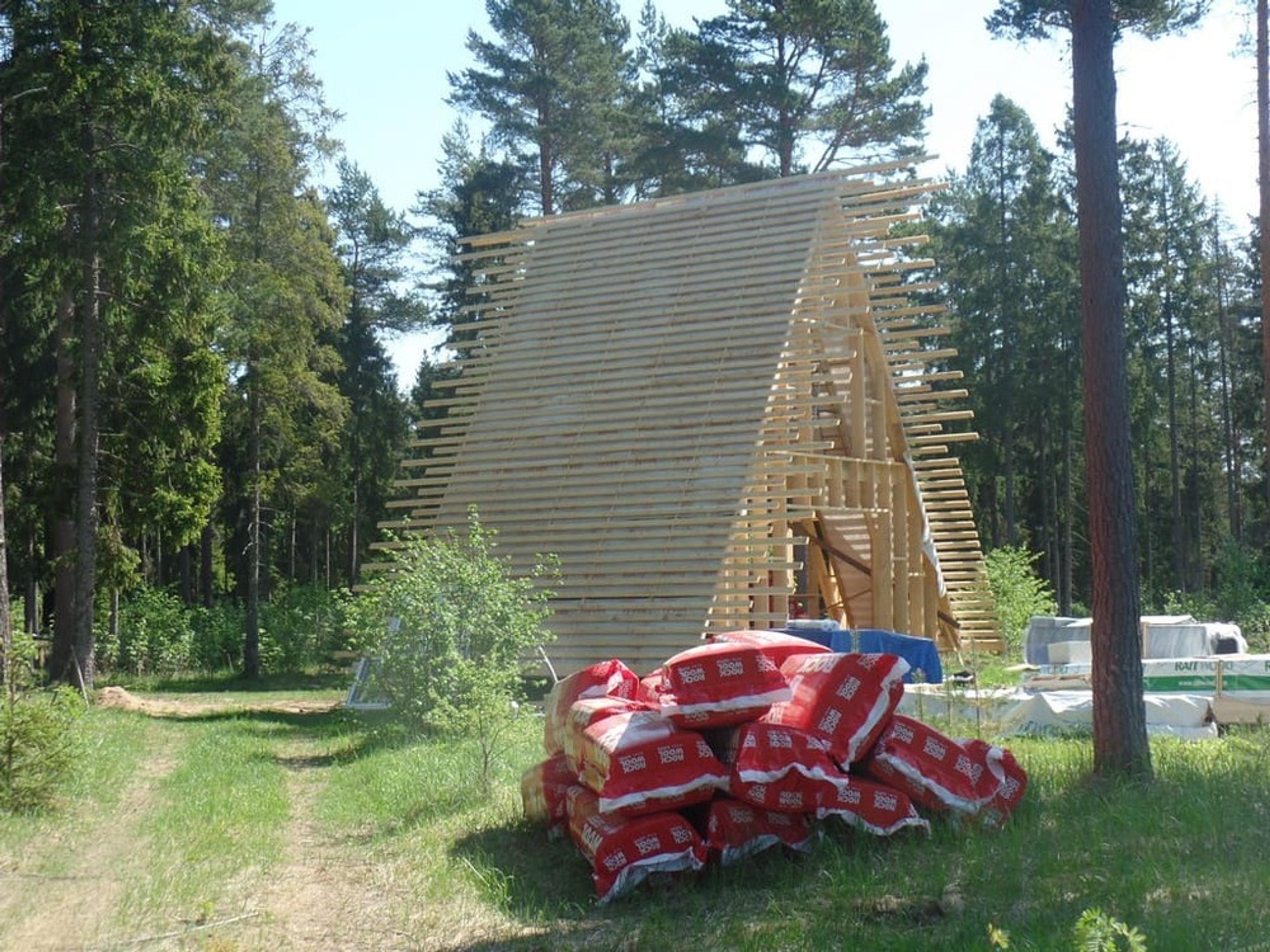 A wooden A-frame skeletal structure with numerous horizontal battens is under construction in a forest, with several red bags of Rockwool insulation piled in the foreground.