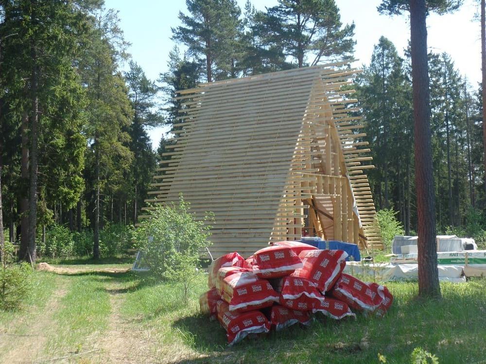A wooden A-frame skeletal structure with numerous horizontal battens is under construction in a forest, with several red bags of Rockwool insulation piled in the foreground.