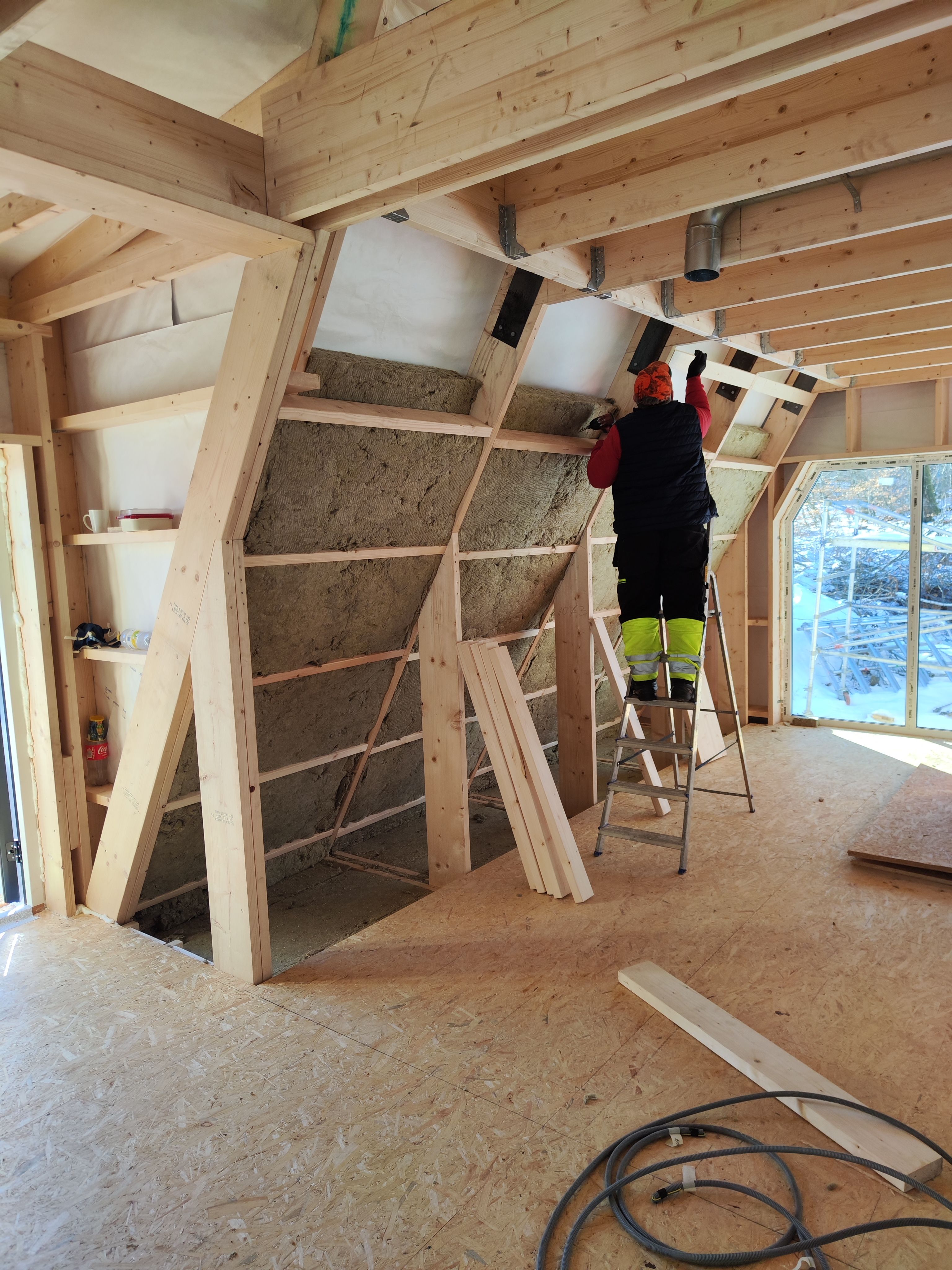 An interior view of an Avrame self-build kit home under construction. A worker standing on a stepladder installs thick batts of insulation into the exposed timber framing of the sloping A-frame wall, showcasing the ease of the internal build process and energy-efficient design.