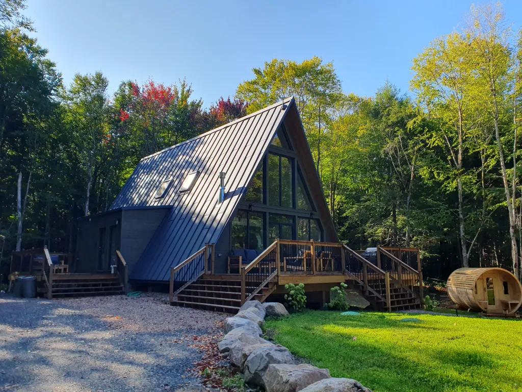 A large, modern A-frame house with a dark metal roof and an expansive wooden deck sits in a sunlit forest clearing next to a barrel sauna.