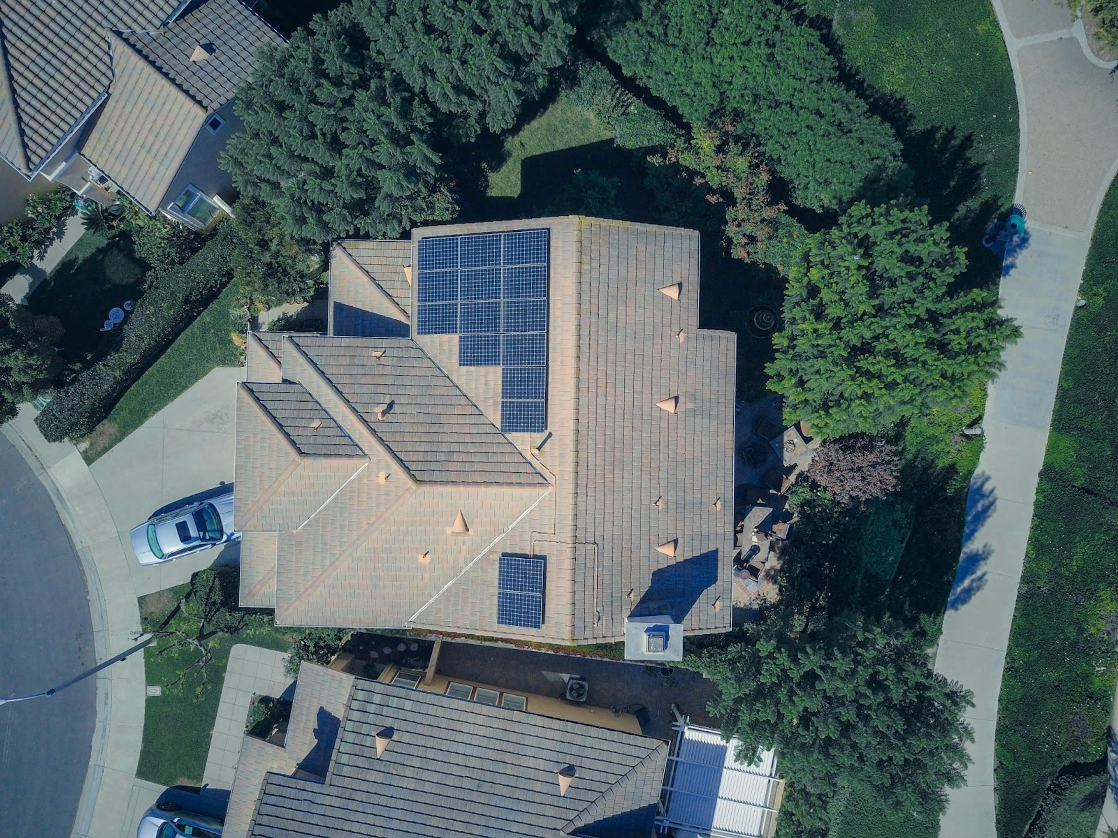 An aerial top-down view shows a suburban house with a grey tiled roof featuring several solar panel arrays installed on different sections of the roofline.