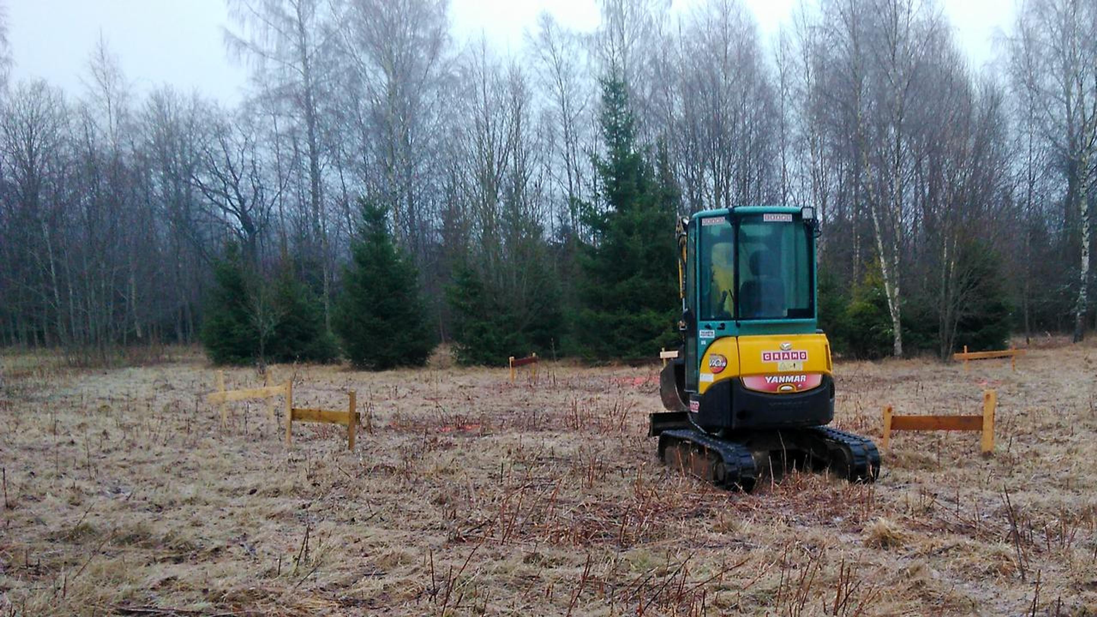 A small yellow excavator sits on a grassy field marked with wooden batter boards, ready to begin groundworks for a new construction project.