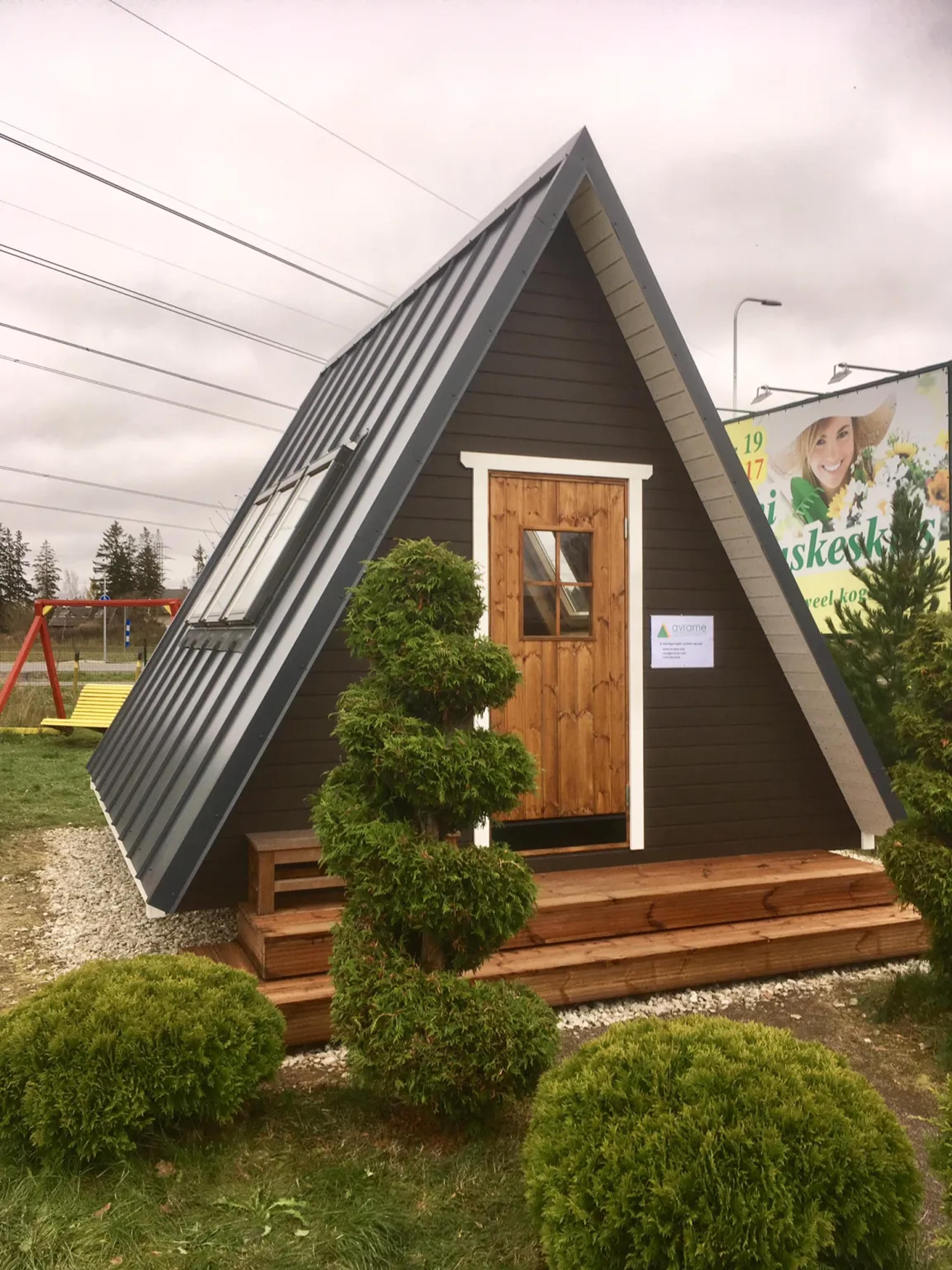 A small, dark-brown Avrame Solo series A-frame cabin sits on a wooden deck surrounded by manicured shrubs under an overcast sky.