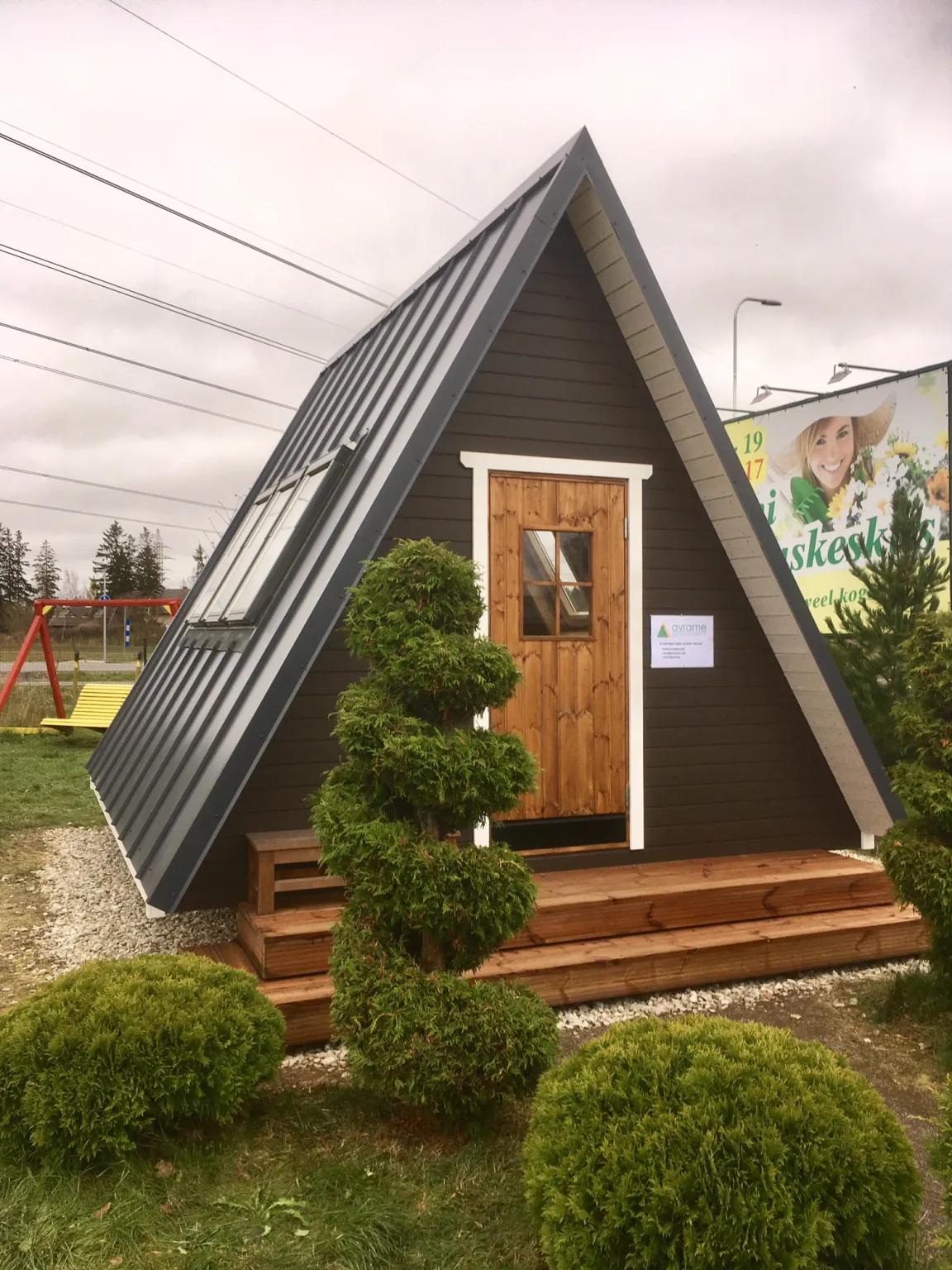 A small, dark-brown Avrame Solo series A-frame cabin sits on a wooden deck surrounded by manicured shrubs under an overcast sky.