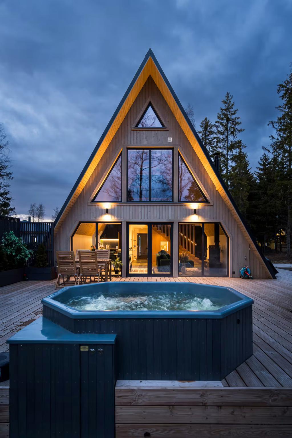 A symmetrical, warmly lit A-frame cabin with large glass windows overlooks a wooden deck featuring a steaming hot tub and a dining area under a twilight sky.