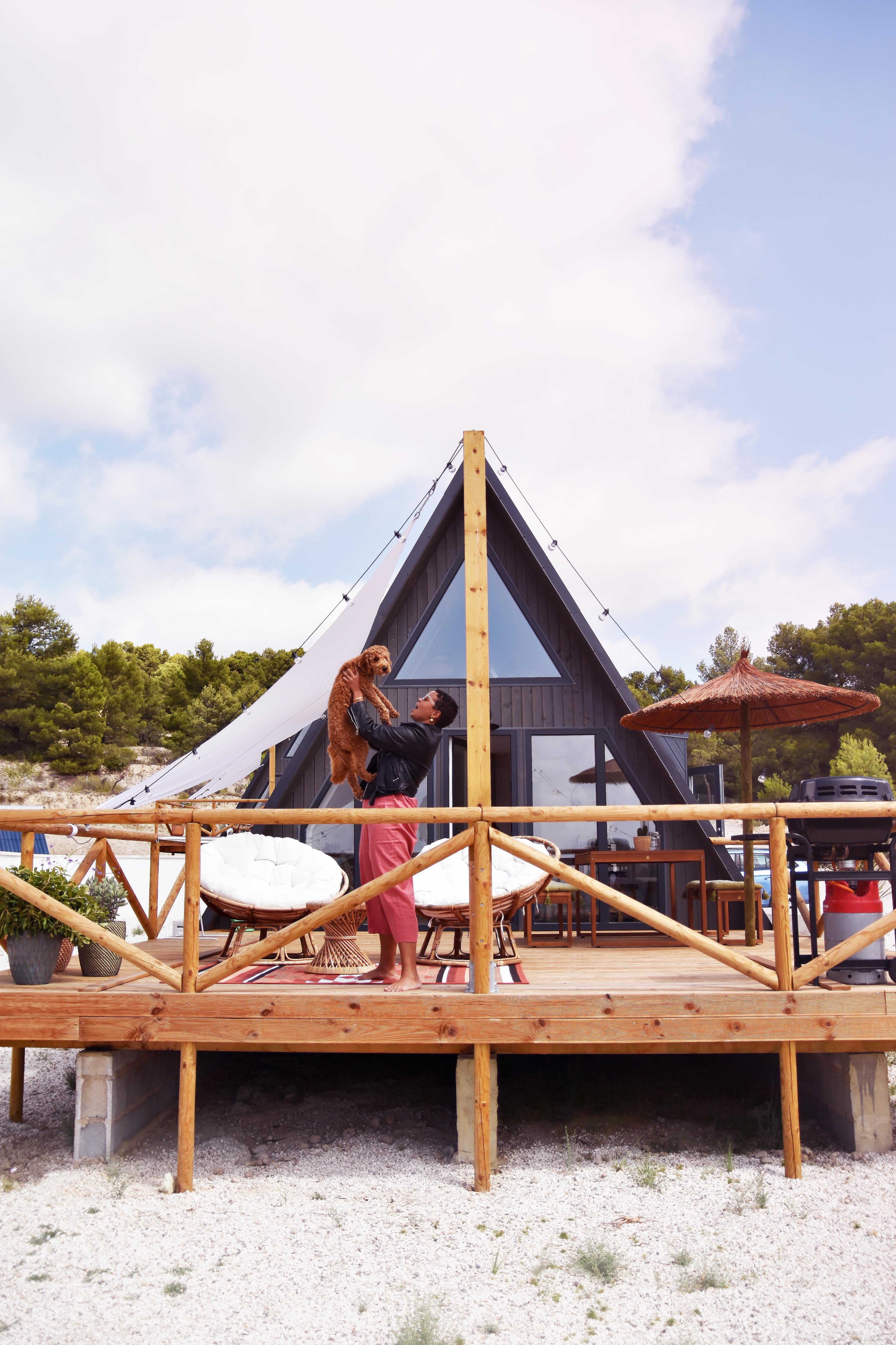 A person stands on the elevated wooden deck of a modern, dark grey A-frame cabin, playfully holding up a fluffy brown dog. The spacious sun deck is styled with white round papasan chairs, a straw umbrella, a barbecue grill, and a white sun sail, set against a backdrop of a clear sky and lush green trees.