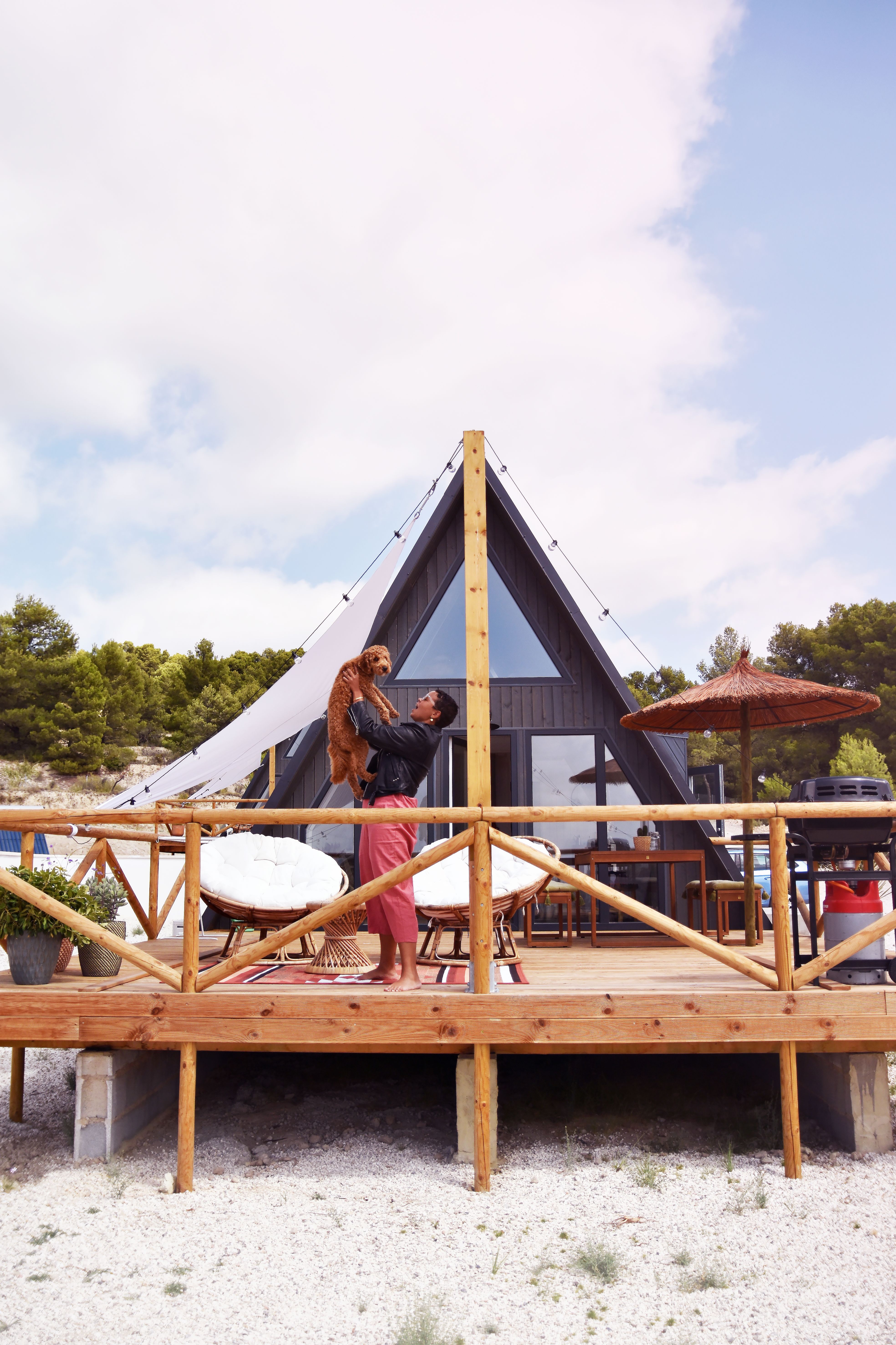 A person stands on the elevated wooden deck of a modern, dark grey A-frame cabin, playfully holding up a fluffy brown dog. The spacious sun deck is styled with white round papasan chairs, a straw umbrella, a barbecue grill, and a white sun sail, set against a backdrop of a clear sky and lush green trees.