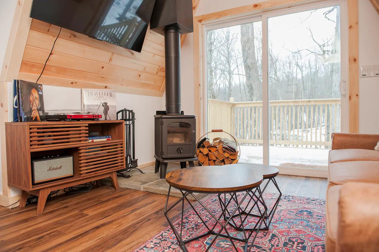 A cozy A-frame living room features a wood-burning stove and a mid-century modern media console next to sliding glass doors that open onto a snowy deck.