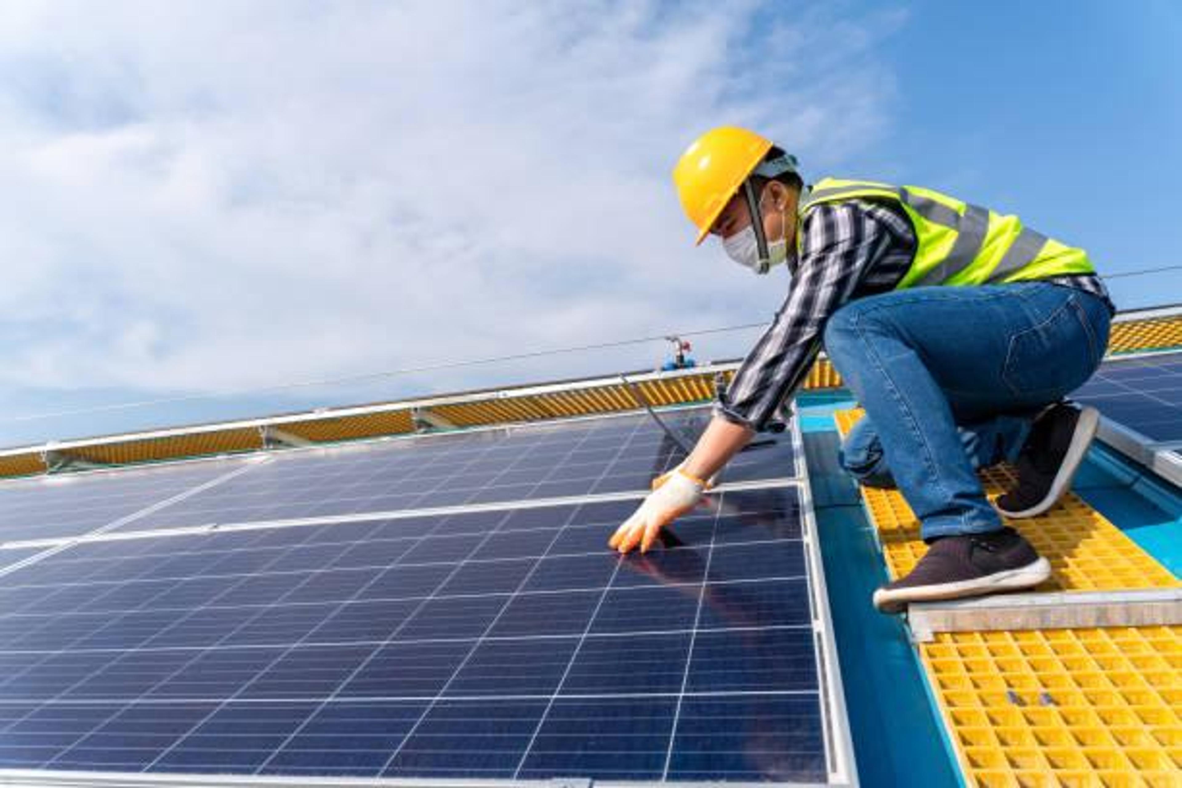 A worker wearing a yellow hard hat, safety vest, and face mask kneels on a rooftop walkway to inspect or install a solar panel.