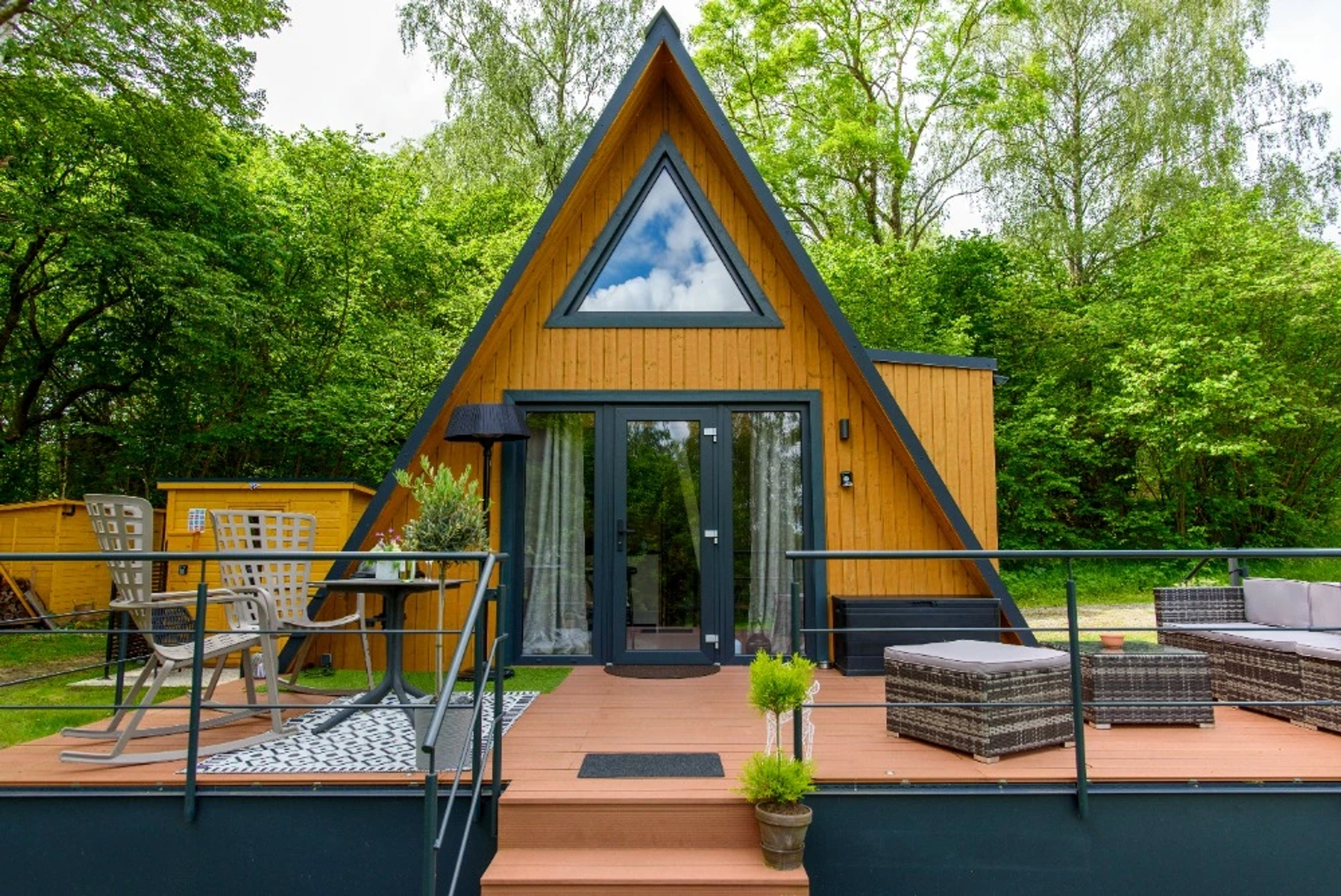 A light wood A-frame house with dark grey trim features a triangular upper window and a spacious elevated deck furnished with rocking chairs, a dining table, and a sectional sofa.