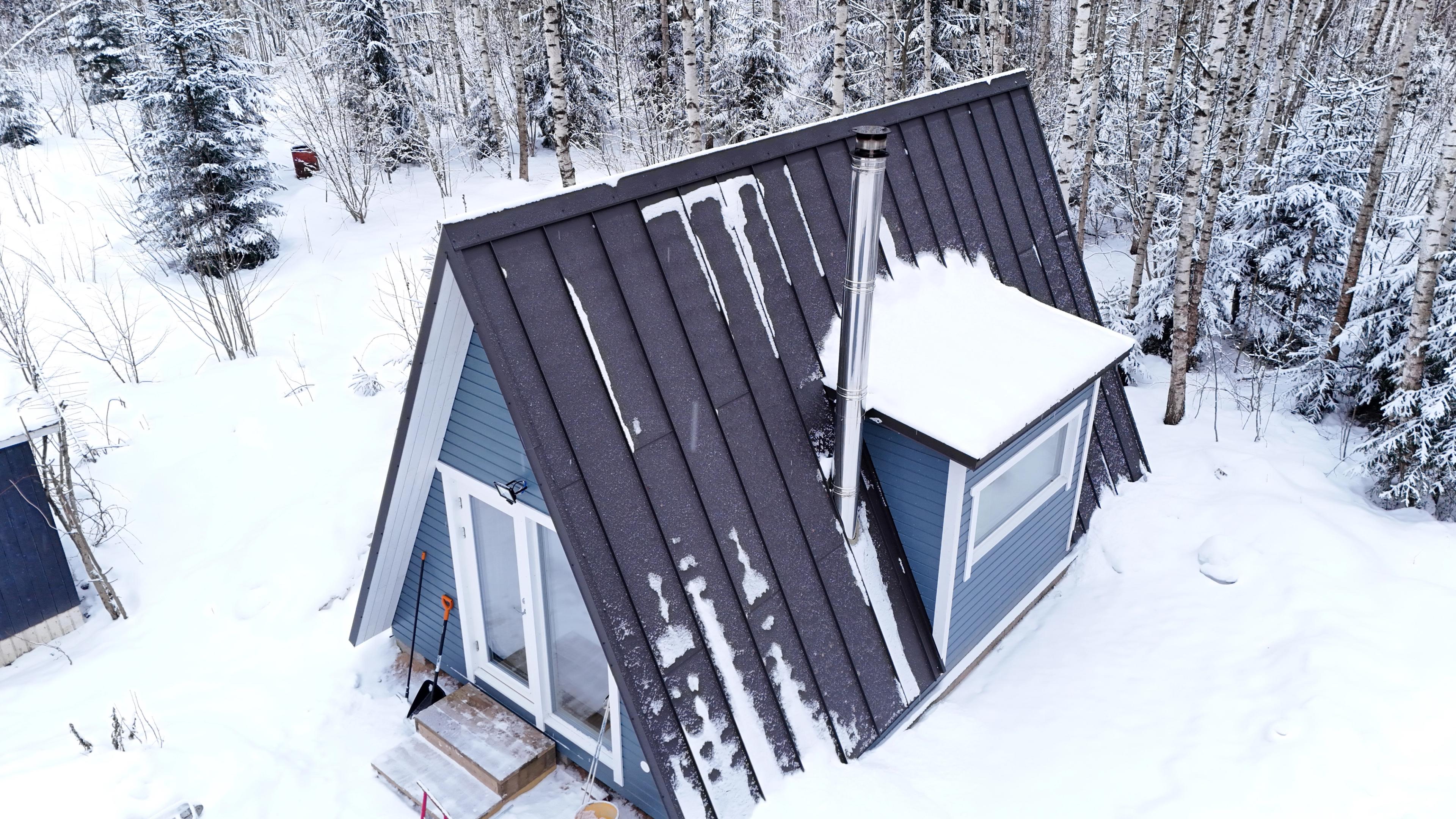An aerial view shows a blue A-frame cabin with a dark metal roof partially covered in snow, featuring a side dormer and a tall silver chimney in a wintry forest.
