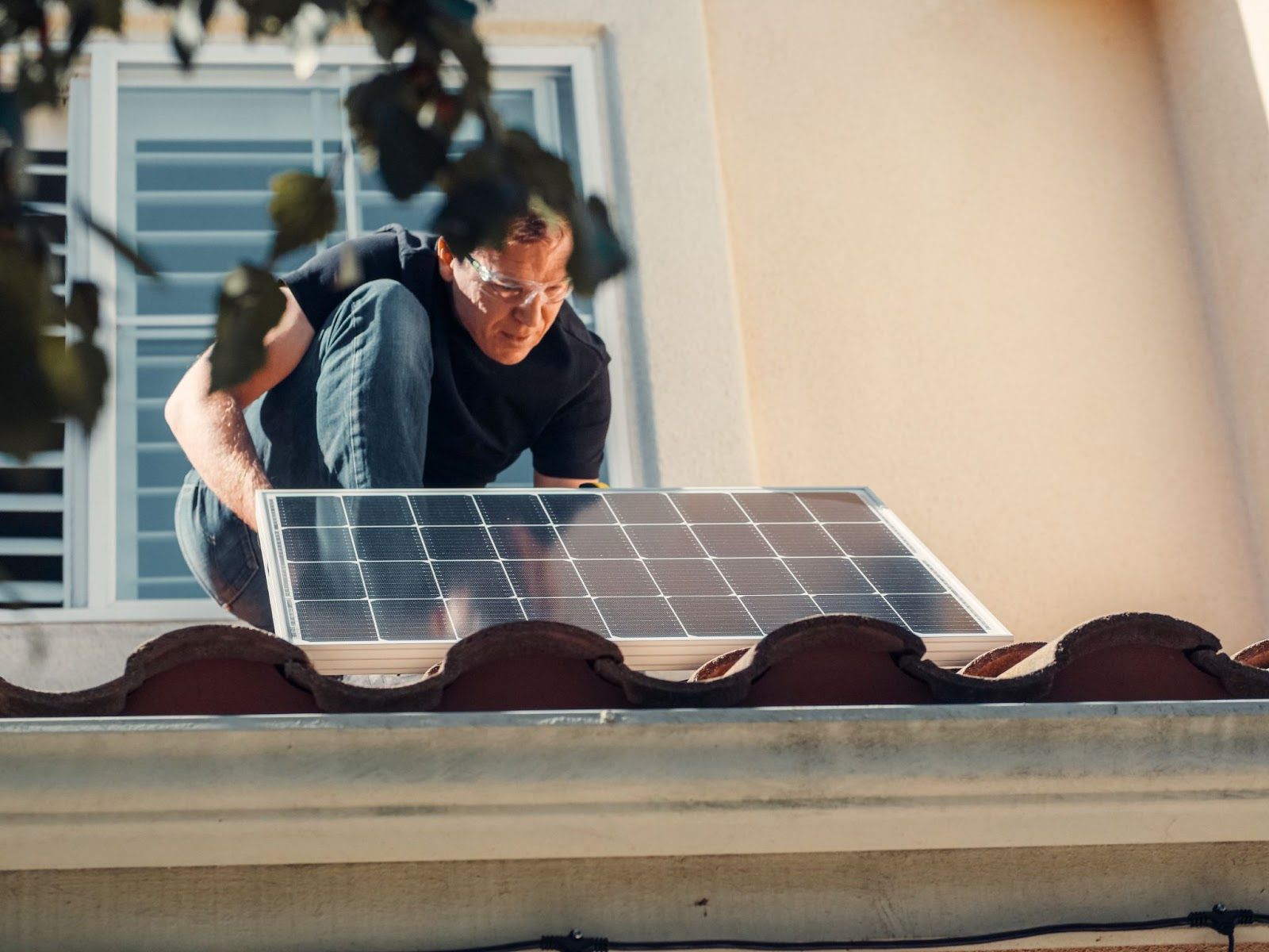 A man wearing protective eyewear kneels on a terracotta tile roof to install a solar panel near a house window.