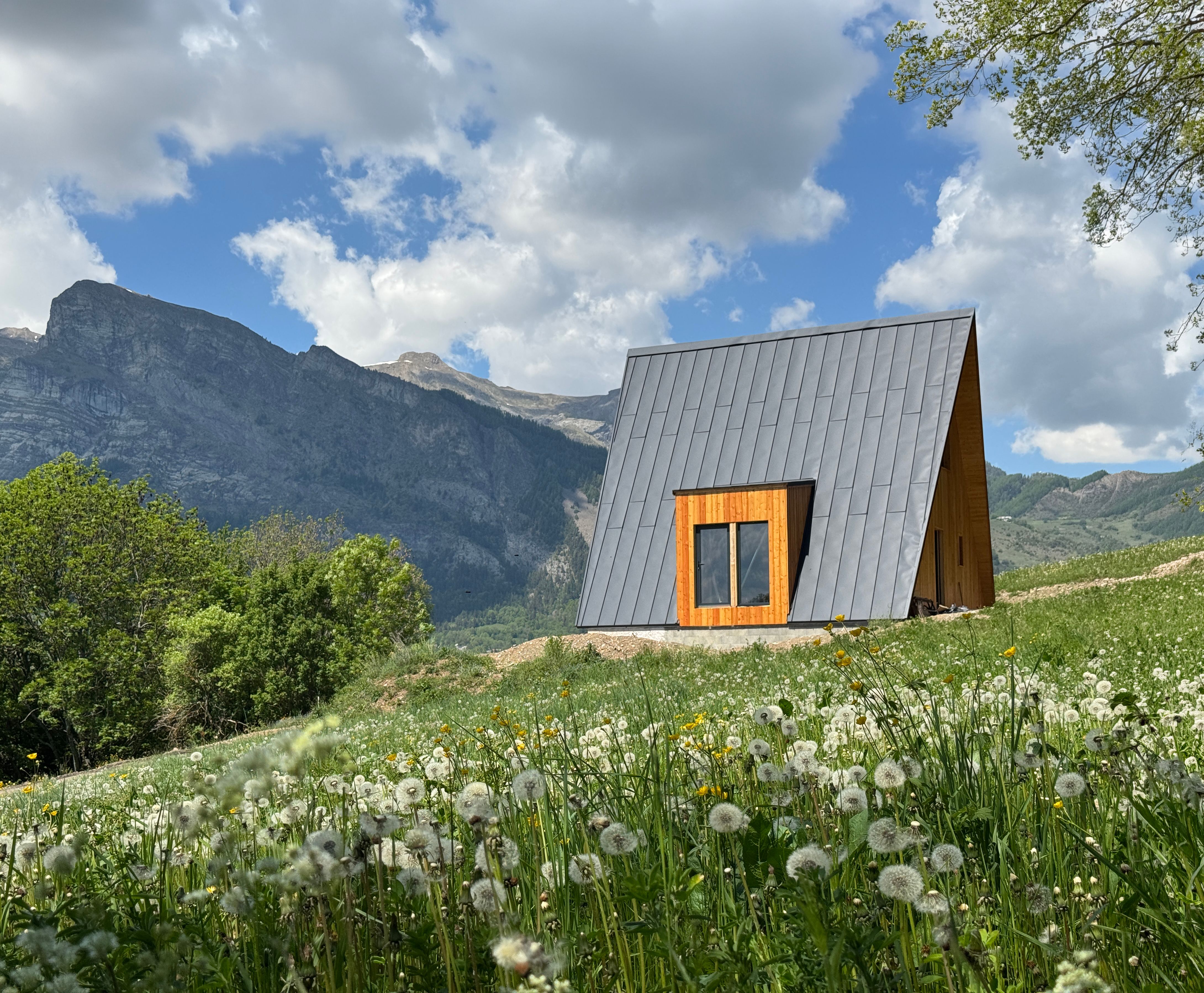 Avrame A-frame house with grey steel roof and timber cladding set in an alpine meadow in southern France, with wildflowers in the foreground and dramatic mountain peaks behind