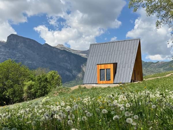 Avrame A-frame house with grey steel roof and timber cladding set in an alpine meadow in southern France, with wildflowers in the foreground and dramatic mountain peaks behind